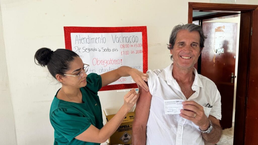 A imagem mostra uma cena em um ambiente clínico ou de saúde pública, onde uma profissional de saúde está aplicando uma vacina em um homem de meia-idade. A profissional usa um uniforme verde e tem os cabelos presos, enquanto o homem veste uma camisa branca e sorri levemente para a câmera. Ele segura um cartão de vacinação na mão esquerda. Ao fundo, há uma placa vermelha com informações em português que indicam o horário de atendimento para vacinação, de segunda a sexta-feira, das 08:00 às 15:00 horas e das 17:00 às 20:00 horas, com a palavra "Obrigatório!" destacada. Também é possível ver um recipiente amarelo para descarte de material biológico, identificado com a marca "EXPELL". A iluminação é clara e o ambiente parece organizado, transmitindo uma atmosfera de cuidado e atenção à saúde.