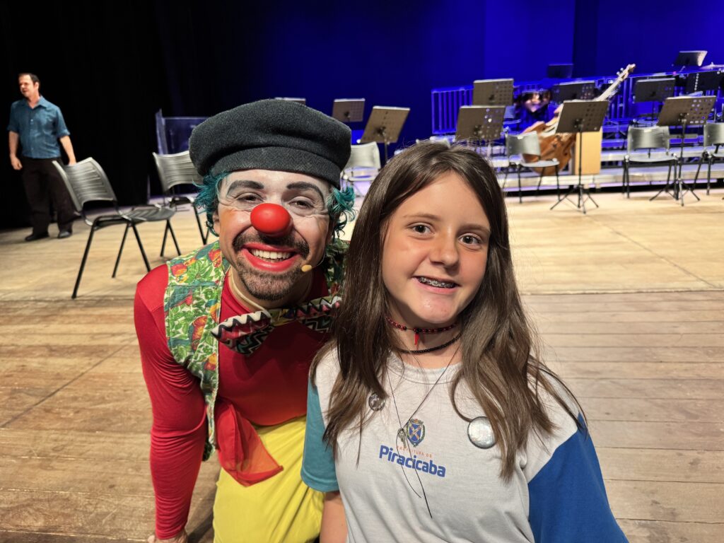 A imagem mostra uma garota jovem e um palhaço posando juntos em um palco de madeira. A garota está mais próxima da câmera, sorrindo e olhando diretamente para ela. Ela tem cabelos castanhos longos, usa uma camiseta branca com mangas azuis, um colar preto com detalhes vermelhos e dois colares com pingentes. O palhaço está ao lado dela, também olhando para a câmera, com um sorriso pintado no rosto, nariz vermelho, cabelo colorido azul e verde, e usa uma boina preta, camisa vermelha, colete estampado com aves tropicais e uma gravata borboleta. Ao fundo, há várias cadeiras e suportes para partituras, indicando que o local é um palco para apresentações musicais. Algumas pessoas aparecem ao fundo, sugerindo que o evento está acontecendo em um ambiente público. A iluminação é quente, destacando os dois personagens principais. No canto superior esquerdo da imagem, aparece o texto "PREFEITURA DE Piracicaba".
