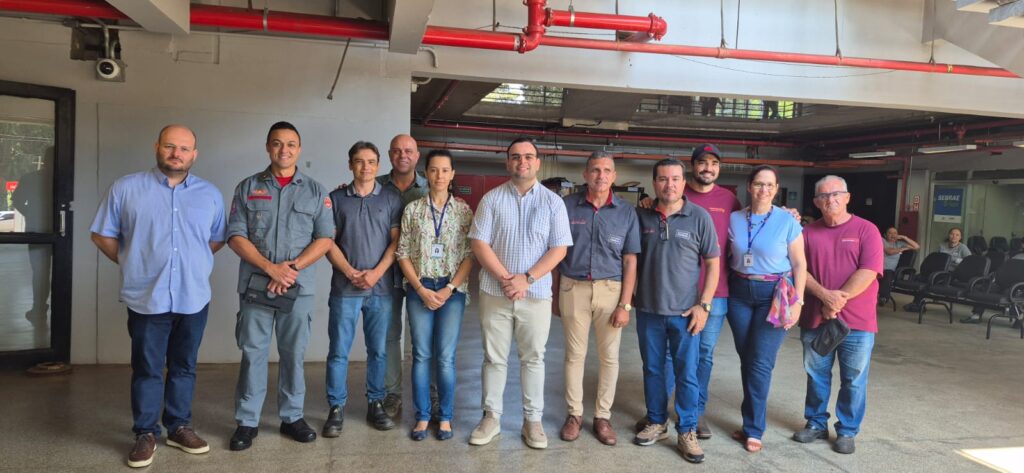 A imagem mostra um grupo de onze pessoas reunidas em um ambiente interno bem iluminado, que parece ser parte de um prédio institucional ou corporativo. Elas estão alinhadas lado a lado, olhando para a câmera, e vestem diferentes tipos de roupas: alguns em trajes casuais ou sociais, enquanto um deles aparece em uniforme do Corpo de Bombeiros, com detalhes em vermelho.