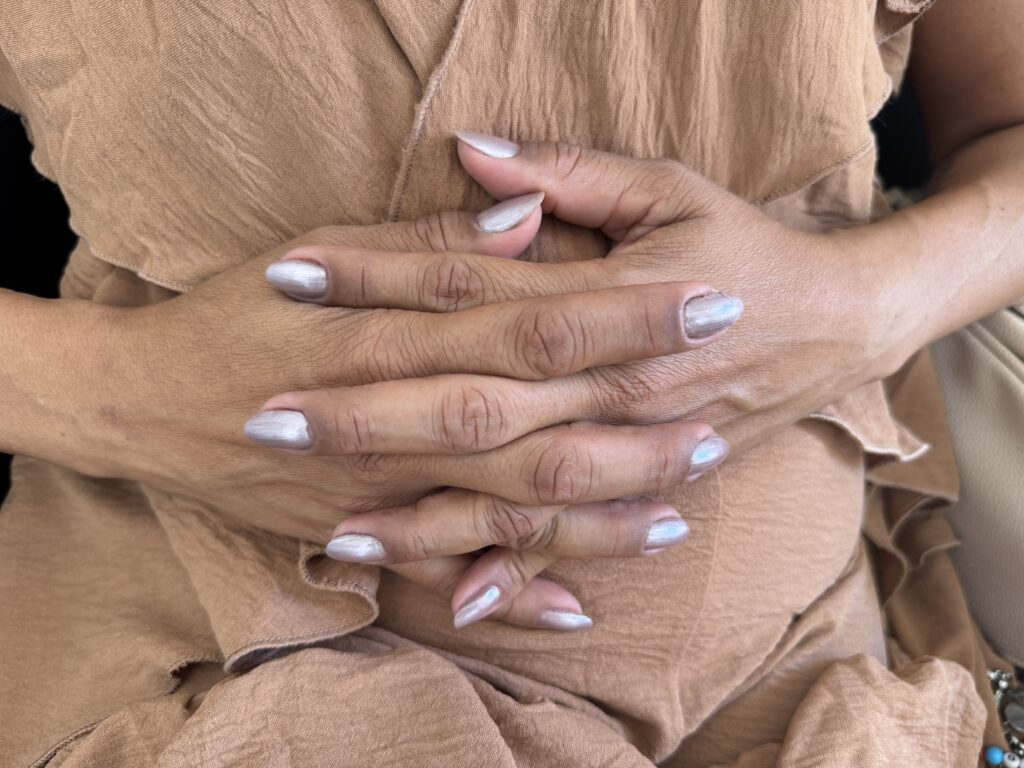 A imagem mostra um close das mãos de uma pessoa, entrelaçadas à frente do corpo. As mãos têm pele de tom médio a escuro, com detalhes visíveis nas linhas e vincos dos dedos e articulações. As unhas estão pintadas com um esmalte perolado, prateado com um brilho iridescente que reflete tons sutis de azul e rosa. A pessoa veste um tecido leve e texturizado, possivelmente de algodão ou linho, em tom bege claro com detalhes em babados. A luz suave destaca a textura da pele e o brilho das unhas, criando uma atmosfera tranquila e contemplativa. A composição é focada nas mãos, com o fundo desfocado, e a imagem foi capturada de um ângulo ligeiramente superior.