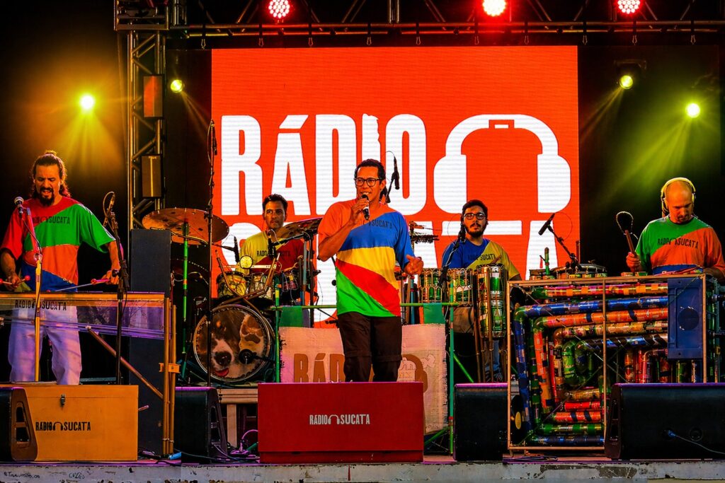 A imagem mostra uma apresentação musical ao vivo de uma banda chamada "Rádio Sucata". O palco está iluminado com luzes coloridas, principalmente amarelas, vermelhas e verdes, criando um ambiente vibrante e animado. Ao fundo, há uma grande tela laranja com o nome da banda "RÁDIO SUCATA" em letras brancas, acompanhada por um desenho de fones de ouvido. No palco, cinco músicos estão posicionados em fila, cada um com seu instrumento. Da esquerda para a direita: o primeiro toca um instrumento de teclas semelhante a um xilofone e veste uma camisa colorida com padrões em verde, vermelho, azul e amarelo; o segundo está na bateria, usando uma camiseta amarela com o nome da banda; o terceiro, que está no centro e é o vocalista, segura um microfone e veste uma camisa bastante colorida com vários tons; o quarto músico toca um instrumento feito com latas e tubos coloridos, usando uma camiseta azul; e o quinto, que está à direita, usa fones de ouvido e toca uma superfície com baquetas, vestindo uma camiseta verde com detalhes em laranja. No palco também há caixas de madeira com o nome "RÁDIO SUCATA" e outros equipamentos de som. A iluminação e as cores vivas das roupas e do cenário conferem um clima festivo e alegre à cena. A banda parece estar realizando uma apresentação energética e criativa, possivelmente com instrumentos não convencionais, reforçando a identidade única do grupo.
