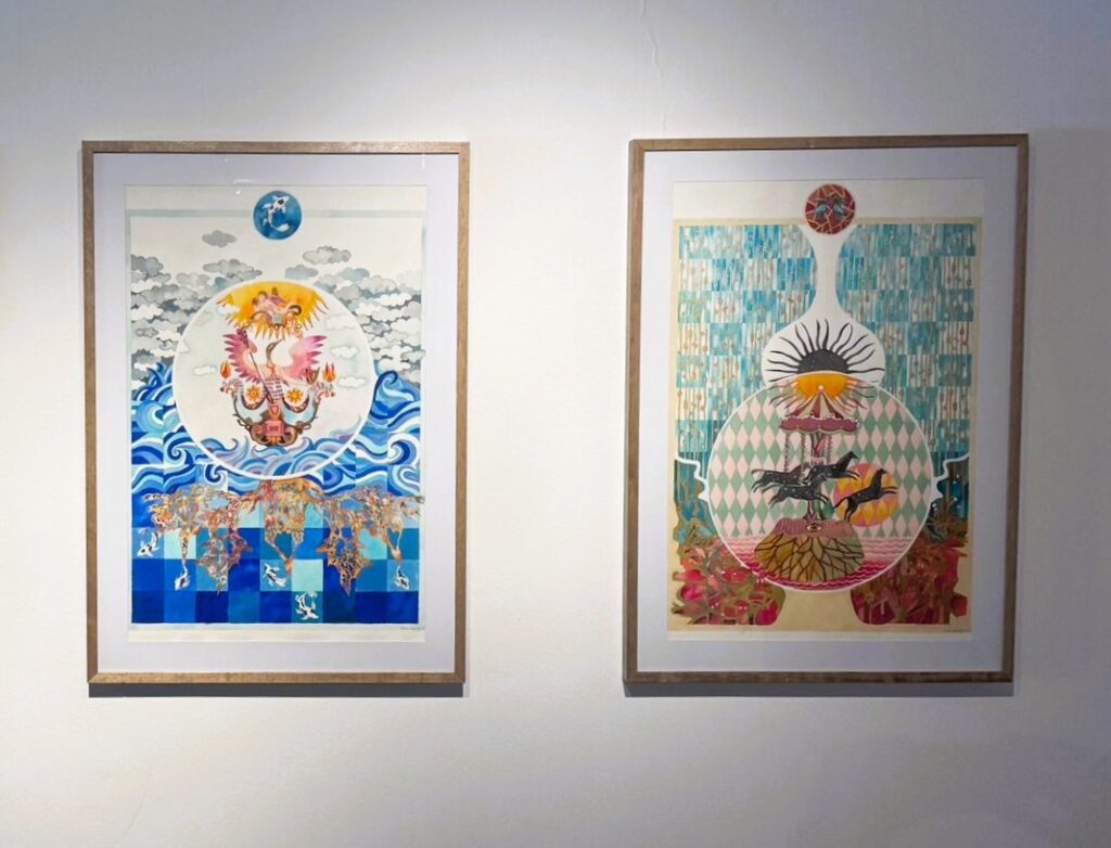 A imagem mostra duas obras de arte penduradas lado a lado em uma parede branca. Ambas são quadros verticais com molduras simples de madeira clara. Quadro da esquerda: Apresenta um círculo central contendo um pássaro semelhante a uma fênix, em tons vibrantes de rosa e laranja, pousado sobre uma estrutura decorativa detalhada. O fundo dentro do círculo é claro, destacando o pássaro. Abaixo, há uma representação de água em tons de azul que se mistura com um padrão quadriculado, além de formas que lembram raízes ou árvores estilizadas. No topo, nuvens cinzas suaves aparecem contra um céu pálido, com um pequeno círculo azul contendo uma forma branca no alto. Quadro da direita: Também tem um círculo central, que mostra um carrossel com três cavalos negros em movimento, diante de um sol estilizado com raios dourados. Abaixo do carrossel, há uma estrutura com um símbolo de olho. O círculo é cercado por um padrão de losangos em verde e rosa. O fundo é composto por linhas verticais azuis e douradas. Acima do círculo, uma forma que lembra um vaso alto termina em um pequeno círculo com dois pássaros. Ambas as obras possuem um estilo surreal e simbólico, com uso intenso de cores vivas e padrões detalhados, transmitindo uma atmosfera onírica e cheia de significado.