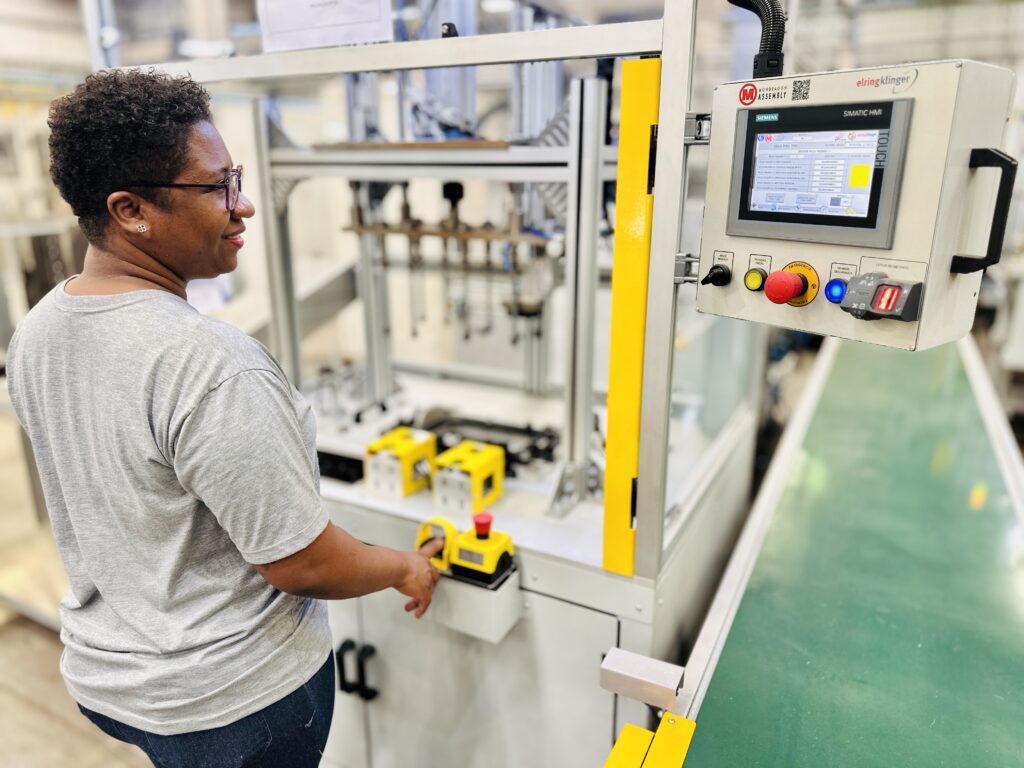 A imagem mostra uma mulher negra de cabelo curto e encaracolado, usando óculos, camiseta cinza e calça jeans azul, em um ambiente industrial, possivelmente uma fábrica. Ela está de pé ao lado de uma máquina grande, apoiando a mão direita em um painel de controle. O painel possui uma tela touchscreen com várias informações em português, incluindo termos como "CICLO POKA YOKE", "PEÇA PADRÃO", "INSERIR PEÇA PADRÃO 1" e a data do último teste "29/4/2026". Também há botões, incluindo um botão de emergência vermelho e um leitor biométrico, indicando medidas de segurança. A mulher parece estar operando ou monitorando a máquina, observando o painel com uma leve expressão de satisfação. O ambiente é bem iluminado, com o equipamento industrial em destaque ao fundo. No painel, aparecem as marcas "MONDRAGON ASSEMBLY", "SIEMENS" e "elringklinger", sugerindo os fabricantes ou fornecedores do equipamento.