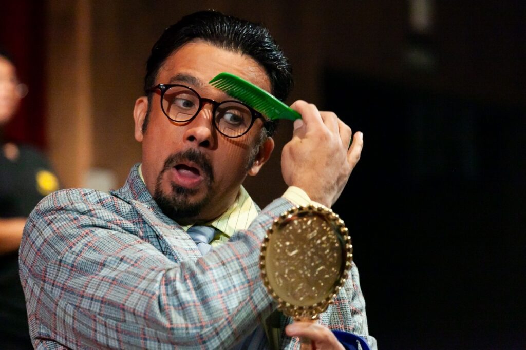 A imagem mostra um homem com cabelo escuro penteado para trás, usando óculos redondos de aro escuro e com um cavanhaque. Ele está segurando um pente verde em uma das mãos, passando-o pelo cabelo, e na outra mão segura um espelho pequeno e dourado, olhando para o espelho com uma expressão de surpresa ou preocupação. Ele veste uma camisa clara, uma camiseta amarela por baixo e um casaco xadrez com predominância de azul, branco e vermelho. O fundo está escuro e desfocado, destacando o rosto e as ações do homem. A iluminação está focada nele, realçando sua expressão facial e os objetos que segura.