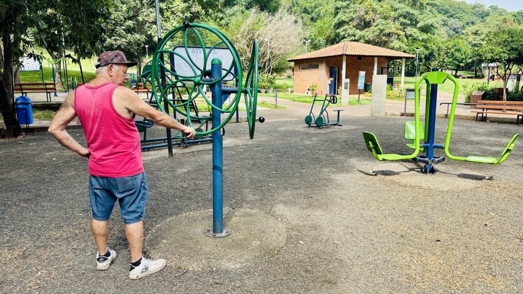 A imagem mostra uma área de exercícios ao ar livre em um parque. No centro da imagem, há um homem idoso de costas, vestindo uma regata rosa e shorts jeans. Ele está em frente a um aparelho de ginástica, que possui duas grandes rodas verdes, e está com a mão esquerda apoiada em uma delas, enquanto a mão direita está na cintura. O aparelho é azul com partes verdes e está fixado em uma base de concreto. Ao fundo, é possível ver outras máquinas de exercícios, bancos e árvores, além de uma pequena construção de tijolos com telhado marrom. O chão é coberto por cascalho e a iluminação indica que é durante o dia, provavelmente no final da manhã ou início da tarde, com sombras suaves. A cena transmite uma atmosfera tranquila de lazer e atividade física ao ar livre, com cores naturais predominantes e um toque vibrante dado pela camisa rosa do homem. A perspectiva da foto é ligeiramente de baixo para cima, destacando o homem e o equipamento de ginástica.
