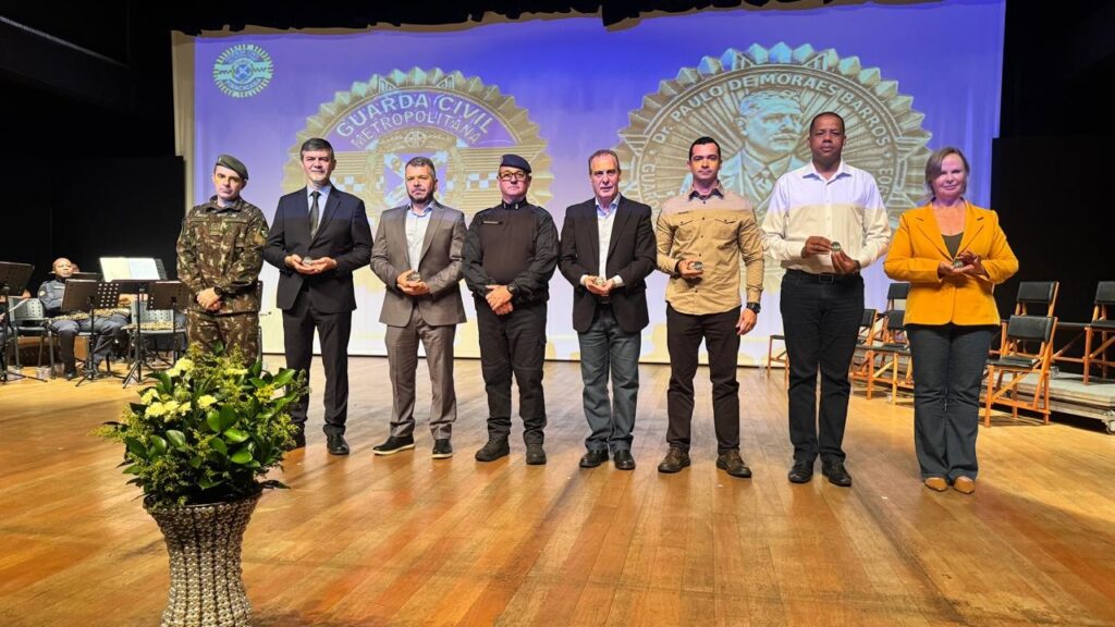 A imagem mostra uma cerimônia formal realizada em um palco, onde oito pessoas estão alinhadas, de frente para a câmera. A maioria dos indivíduos está vestida com roupas formais, como ternos e uniformes militares. O primeiro homem à esquerda usa um uniforme camuflado e um boina, sugerindo que faz parte de uma força de segurança ou militar. Os outros seis homens e a mulher, que veste um blazer amarelo, seguram pequenos objetos que parecem ser medalhas ou troféus, indicando que estão sendo homenageados ou premiados. Ao fundo, há um grande painel azul com um emblema circular contendo o texto "GUARDA CIVIL METROPOLITANA" e a imagem de uma pessoa, provavelmente uma figura importante ou homenageada. Ao lado, há o nome "DR. PAULO DE MORAES BARROS", possivelmente o nome do evento, da instituição ou de um homenageado. Na parte frontal do palco, à esquerda, está um vaso grande com arranjos florais em branco e amarelo, que adiciona um toque decorativo ao ambiente. Também é possível ver uma pequena orquestra sentada ao fundo, com músicos tocando instrumentos, o que sugere que a cerimônia conta com apresentação musical. O cenário é iluminado de forma clara, destacando os participantes da cerimônia e o painel ao fundo. O ambiente é formal e solene, típico de eventos oficiais de reconhecimento ou premiação.
