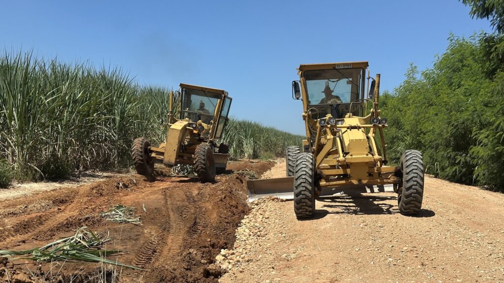 A imagem mostra uma cena rural onde dois motoniveladoras amarelas estão trabalhando em uma estrada de terra ao lado de uma plantação. A motoniveladora em primeiro plano está sobre uma estrada de cascalho, enquanto a segunda está nivelando o solo ao lado, próximo a uma plantação de cana-de-açúcar. O céu está claro e azul, indicando um dia ensolarado. Há vegetação verde ao redor, incluindo árvores e a plantação alta de cana. As máquinas parecem estar realizando manutenção ou preparação do terreno, e os operadores são visíveis dentro das cabines. A luz do sol cria sombras definidas, sugerindo que a foto foi tirada durante o dia. A cena transmite uma sensação de trabalho agrícola e infraestrutura rural.