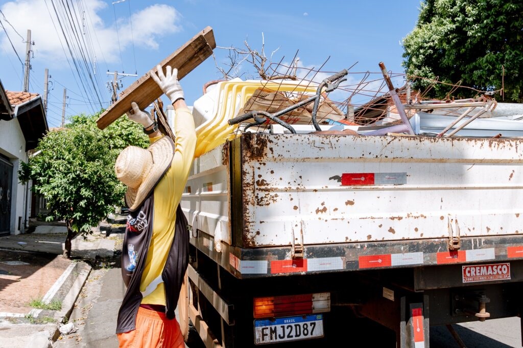 A imagem mostra uma cena ao ar livre, em uma rua residencial durante o dia, com céu azul e algumas nuvens. Em primeiro plano, há uma pessoa vestindo chapéu de palha, camiseta amarela, shorts laranja e luvas brancas, que está carregando um pedaço de madeira para dentro de um caminhão pequeno, tipo caçamba. O caminhão está carregado com vários tipos de entulho, incluindo pedaços de madeira, plásticos, metais e até partes de bicicleta. O caminhão é branco, com sinais de uso, como ferrugem e pintura desgastada. Na lateral e na traseira do veículo, há faixas refletivas vermelhas e brancas, além de adesivos com a marca "CREMASCO" e informações de contato. A placa do caminhão indica que o veículo é do Brasil, com o número "FMJ2D87". Ao fundo, é possível ver casas, árvores e postes com fiação elétrica, reforçando o ambiente residencial. A luz do sol é forte, criando sombras bem definidas na cena, o que sugere que a foto foi tirada no período da manhã ou início da tarde. A imagem transmite a ideia de trabalho manual, possivelmente relacionado à limpeza, reciclagem ou remoção de entulho.