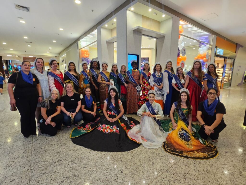 A imagem mostra um grande grupo de mulheres, provavelmente competidoras de um concurso de beleza ou evento cultural, posando para uma foto em ambiente interno, que parece ser um shopping center. Muitas das mulheres usam faixas com nomes de países, como "Suíça", "Tailândia", "Holanda", "Reino Unido", "Alemanha", "Coreia", "Moçambique", "Portugal" e "Brasil", indicando que representam diferentes nações. Algumas estão usando roupas tradicionais coloridas, enquanto outras vestem roupas mais modernas, mas todas parecem estar em traje festivo ou formal. Duas mulheres no centro da frente usam saias longas e coloridas, destacando-se no grupo. A maioria está sorrindo, transmitindo um clima de celebração e alegria. O ambiente é bem iluminado, com piso brilhante e decoração simples, incluindo balões ao fundo. A foto foi tirada de um ângulo levemente superior, capturando toda a diversidade do grupo e o espaço ao redor. A imagem transmite uma sensação de união, diversidade cultural e confraternização entre as participantes.