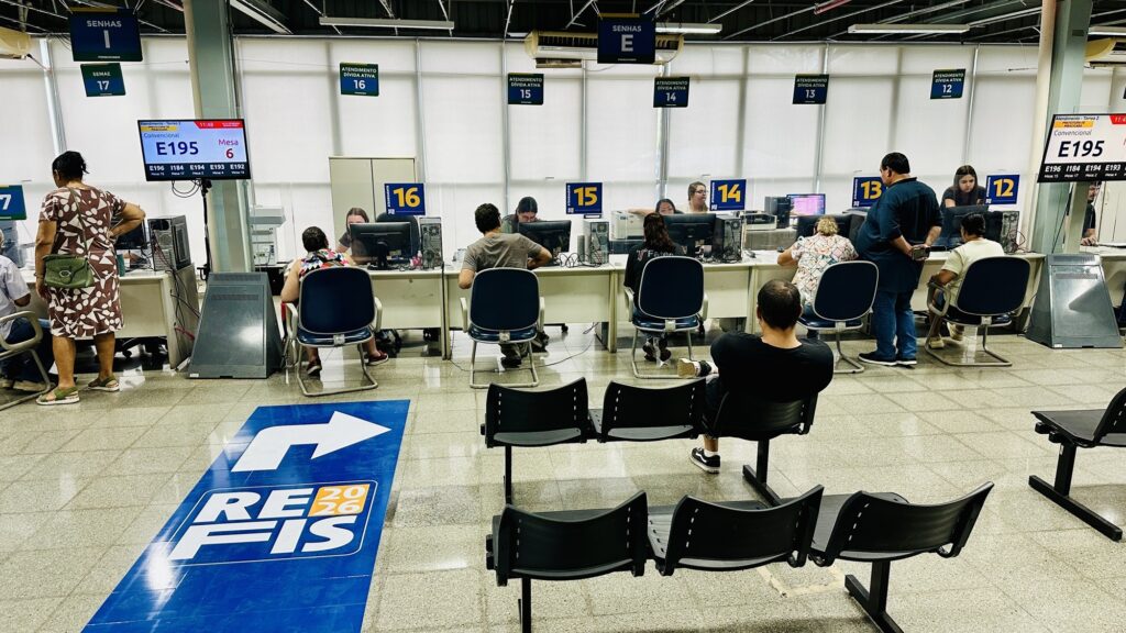 A imagem mostra um ambiente interno de um órgão público, possivelmente uma agência de atendimento para negociação de dívidas, como o REFIS 2026, conforme indicado no adesivo grande no chão com uma seta azul apontando para os guichês. No fundo, há uma fileira de guichês numerados de 12 a 16, onde funcionários estão sentados atrás de computadores atendendo pessoas. Acima dos guichês, painéis digitais exibem senhas e números de atendimento, como "E195" e "Mesa 6". Alguns atendentes estão em pé ou conversando com os clientes. Na parte da frente, há várias cadeiras pretas organizadas em fileiras para espera, com poucas pessoas sentadas. Uma delas é um homem de camiseta azul com as costas para a câmera. Do lado esquerdo, uma mulher está em pé, vestindo um vestido com estampa marrom e branca. O ambiente é iluminado por luz natural e artificial, com janelas grandes cobertas por persianas brancas. O espaço parece limpo, organizado e funcional, projetado para facilitar o fluxo dos cidadãos que aguardam atendimento para regularizar suas dívidas.
