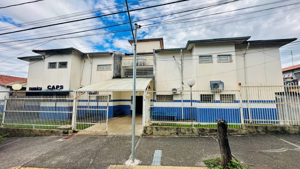 A imagem mostra a fachada de um prédio público de dois andares, identificado como "FARMÁCIA CAPS" (Centro de Atenção Psicossocial). O edifício é branco com uma base revestida de azulejos azuis. Ele está cercado por uma grade metálica branca e possui uma entrada principal com um pequeno toldo também branco. Na frente do prédio, há um poste de iluminação ou de energia elétrica com vários fios cruzando a imagem. A calçada em frente tem piso tátil para acessibilidade, e à direita da imagem há um toco de árvore. O céu está claro com algumas nuvens, indicando um dia ensolarado. O prédio apresenta algumas marcas de desgaste, principalmente próximas ao telhado. Há janelas com grades e aparelhos de ar-condicionado instalados no segundo andar. Uma pessoa está parcialmente visível na entrada, em uma área sombreada. No geral, a imagem transmite a ideia de um serviço público de saúde localizado em uma área urbana, com elementos que indicam acessibilidade e infraestrutura básica.