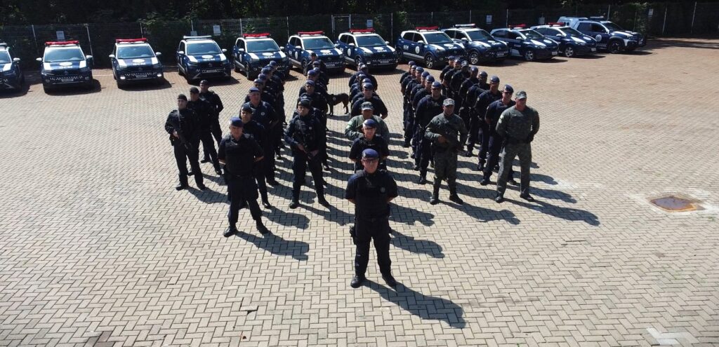 A imagem mostra um grupo de policiais da Guarda Civil Municipal (GCM) reunidos em um pátio externo pavimentado com blocos intertravados em padrão espinha de peixe. Eles estão em formação, alguns em uniformes azul escuro e outros com uniformes camuflados, todos aparentando estar em posição de atenção ou em postura formal. Ao fundo, há várias viaturas policiais da GCM, também na cor azul escuro, com sirenes vermelhas e azuis no teto. O ambiente é ao ar livre, com iluminação forte e sombras definidas, indicando um dia ensolarado. Ao fundo, pode-se ver uma cerca e algumas árvores, sugerindo que o local pode ser uma área de treinamento ou estacionamento oficial da corporação. A cena transmite organização, disciplina e prontidão dos agentes.