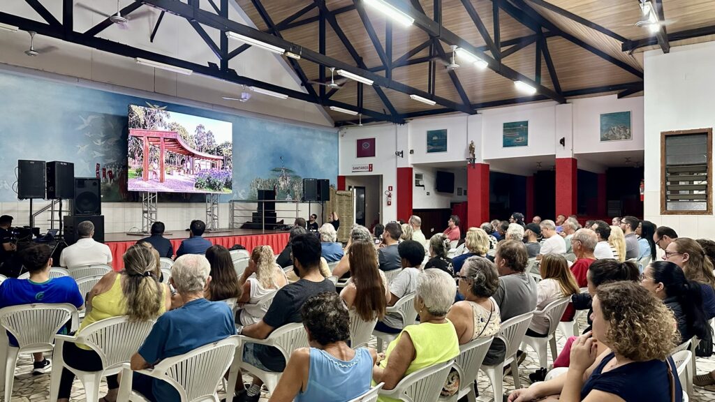 A imagem mostra uma reunião ou apresentação realizada em um espaço interno amplo, com um teto alto e estrutura de madeira escura aparente. Há muitas pessoas sentadas em cadeiras plásticas brancas, todas voltadas para um palco onde está instalada uma grande tela de LED. Na tela, é exibida uma imagem digital de um projeto arquitetônico, provavelmente uma pérgola ou estrutura de madeira para área externa. O ambiente é bem iluminado com luzes fluorescentes no teto. As paredes são claras, com alguns detalhes em vermelho e murais azuis com desenhos de pássaros e paisagens. Ao lado do palco, há caixas de som pretas empilhadas. No canto da imagem, uma placa indica a direção para os sanitários. O público é diversificado, composto por adultos e idosos, e todos parecem atentos à apresentação. A atmosfera sugere um evento comunitário, palestra ou reunião para discussão de um projeto local.