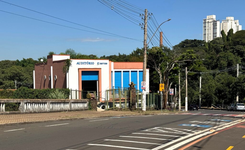 A imagem mostra um prédio branco com detalhes em terracota, identificado como "AUDITÓRIO semae", localizado em uma esquina de rua. O edifício possui janelas com persianas azuis e está cercado por um muro baixo de pedra com uma grade metálica por cima. Na frente do prédio, há uma calçada e uma faixa de pedestres pintada na rua. Próximo ao prédio, há uma placa de estacionamento com símbolo de acessibilidade e uma placa amarela de advertência para pedestres. Ao fundo, vê-se uma área arborizada e, mais distante, prédios residenciais altos. O céu está claro e azul, indicando um dia ensolarado. A cena transmite um ambiente urbano tranquilo e organizado.