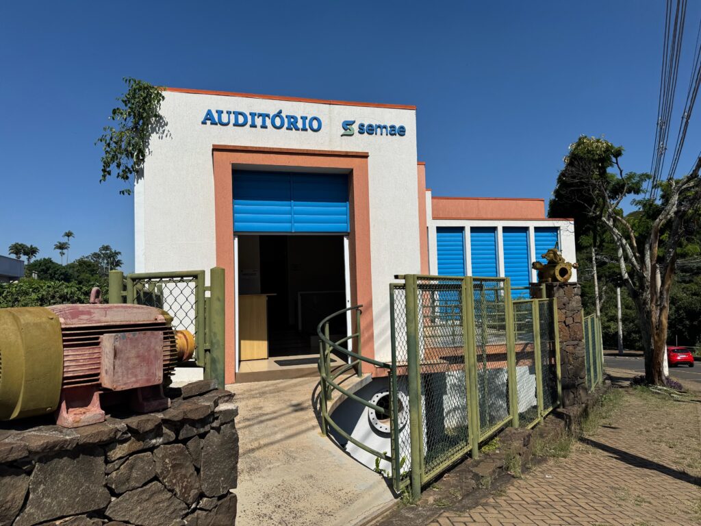 A imagem mostra a fachada de um prédio com a palavra "AUDITÓRIO" escrita em letras grandes e azuis, junto com o logotipo da empresa ou órgão chamado "semae". O prédio é branco com detalhes em laranja, como a faixa superior e a moldura da entrada. A porta de entrada e as janelas laterais são azuis, com um design de venezianas. Na parte da frente, à esquerda, há um grande motor antigo, pintado de vermelho com partes amarelas, apoiado sobre um muro de pedras. À direita da entrada, há uma cerca verde de metal, e um pilar de pedra onde está fixado um equipamento industrial, possivelmente uma válvula ou bomba, pintado de verde claro. O ambiente é externo, com árvores e palmeiras ao fundo, indicando um clima tropical ou subtropical. O céu está limpo e azul, mostrando que é um dia ensolarado. No chão, uma calçada de concreto leva até a entrada, com um corrimão verde curvo ao lado, sugerindo acessibilidade. A imagem transmite a ideia de um espaço público ou institucional, possivelmente ligado a serviços de saneamento ou meio ambiente, dada a presença dos equipamentos industriais e o nome "semae", que pode estar relacionado a esses setores.