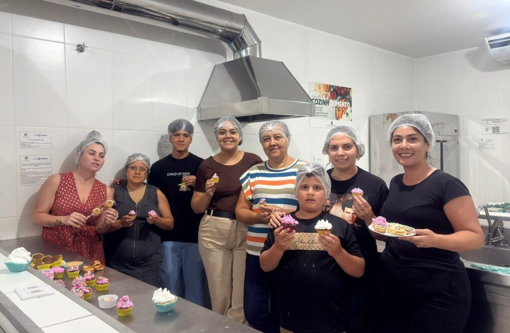 A imagem mostra um grupo de oito pessoas em uma cozinha industrial bem iluminada. Todos usam toucas brancas, indicando cuidados com higiene alimentar. O grupo é composto por adultos e uma criança, que está segurando cupcakes decorados com cobertura rosa e branca. Algumas pessoas estão segurando cupcakes e outras um prato com pequenos doces, possivelmente mini donuts. Ao fundo, há uma parede com azulejos brancos, uma coifa de inox e equipamentos típicos de cozinha, como pia e geladeira. No canto superior direito, há um cartaz com o texto "PROJETO COZINHALIMENTO". A atmosfera geral é de alegria e celebração, sugerindo que o grupo participou de uma atividade culinária, talvez uma oficina ou projeto de aprendizado gastronômico.