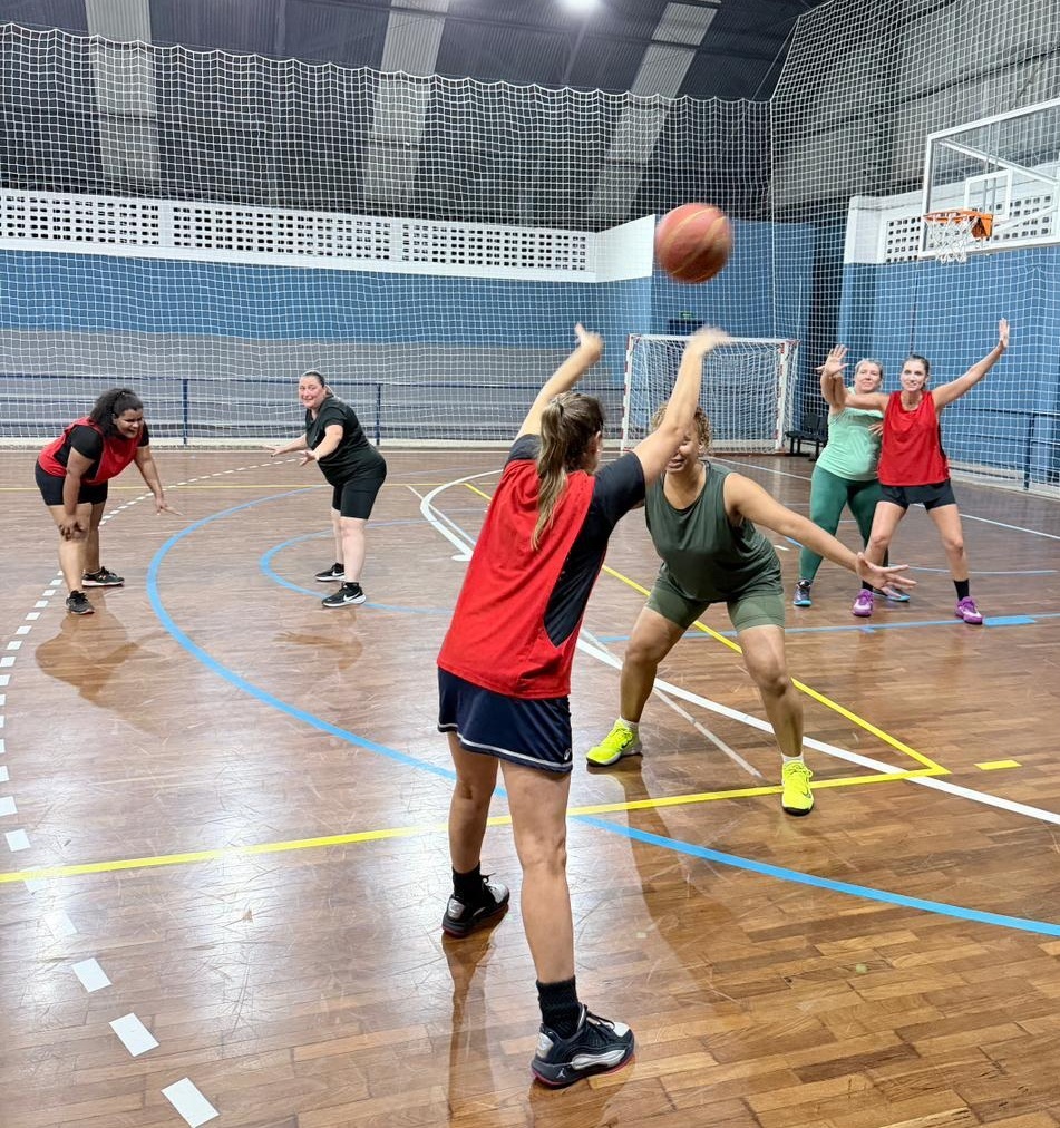 A imagem mostra um grupo de seis mulheres jogando basquete em uma quadra coberta com piso de madeira. O foco principal está em uma bola de basquete no ar, sendo passada de uma jogadora para outra. As mulheres estão vestidas com roupas esportivas, como shorts e regatas ou camisetas, em cores variadas como vermelho, preto, verde e cinza. A quadra possui marcações em azul e amarelo, e ao fundo há uma rede azul e uma cesta de basquete. A iluminação é artificial, vinda de cima, refletindo no piso polido. A expressão das jogadoras é de concentração e envolvimento no jogo. A imagem foi capturada de um ângulo levemente elevado, olhando para baixo, mostrando a ação em andamento.