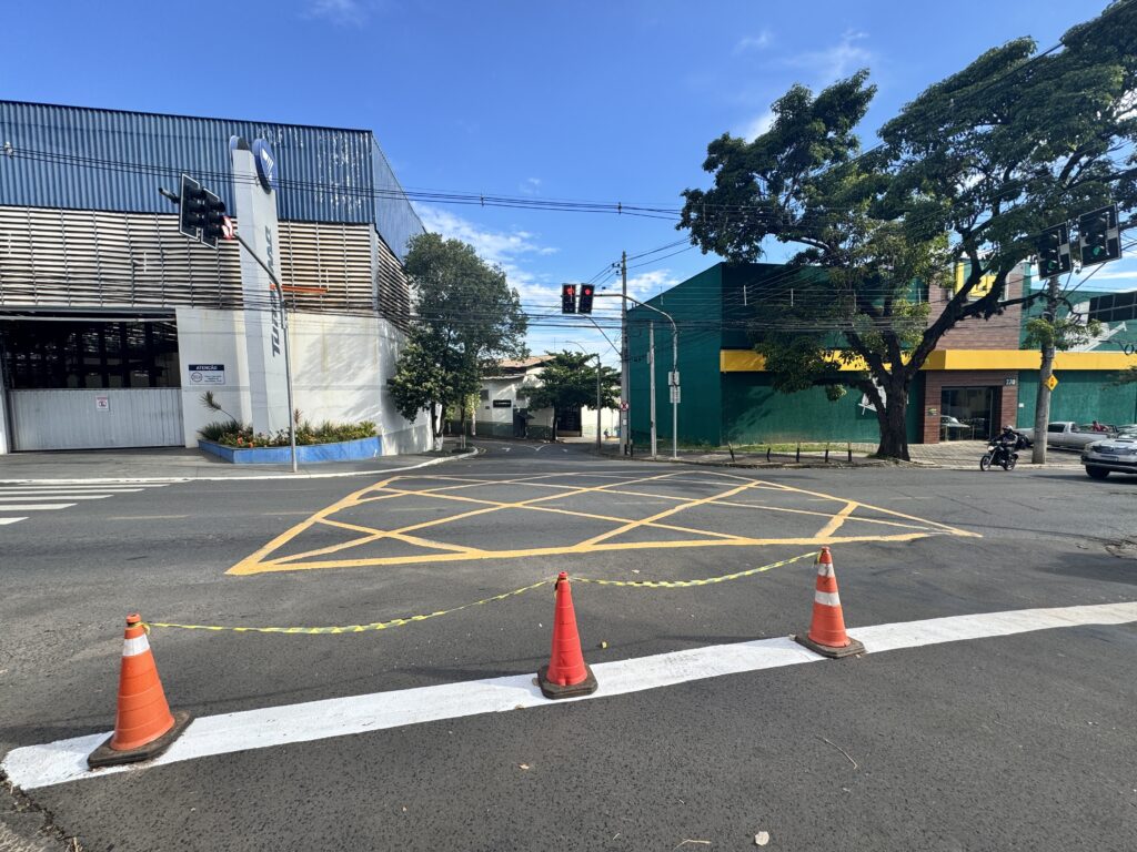 A imagem mostra um cruzamento urbano durante o dia, com um céu claro e algumas nuvens. No primeiro plano, há cones de trânsito laranja conectados por uma fita amarela e preta, posicionados logo após uma linha branca de parada. No chão, no centro do cruzamento, há marcações amarelas em ziguezague que indicam uma área onde os veículos não devem parar, conhecida como "caixa amarela" ou "caixa de retenção". À esquerda, há um edifício grande com estrutura de metal e telhado azul, com uma placa escrita "ATENÇÃO" e um letreiro com o nome "TUBOMAG". À direita, um prédio verde e amarelo com o número "270" visível na fachada. Um motociclista, vestido com roupas escuras e capacete, está à direita da imagem, esperando para seguir no cruzamento. A cena sugere que a área está temporariamente controlada ou em manutenção, devido à presença dos cones e da fita, e o trânsito está organizado para evitar que os veículos avancem até a caixa amarela, respeitando as regras de trânsito locais. A iluminação é clara e destaca bem as cores vibrantes dos cones e das marcações no asfalto.