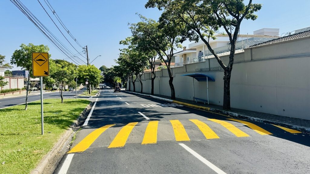 A imagem mostra uma cena urbana durante o dia, com céu claro e ensolarado. A rua principal é asfaltada e possui marcas amarelas no chão, indicando uma lombada para reduzir a velocidade dos veículos. À esquerda da rua, há uma faixa de grama e uma placa de trânsito amarela em formato de losango, que alerta sobre a presença da lombada, com uma seta apontando para baixo. À direita da rua, há uma calçada, um ponto de ônibus com um abrigo azul e um banco, além de um muro alto, claro, com arame farpado no topo. Árvores com folhagem verde estão alinhadas ao longo da calçada, proporcionando sombra. Ao fundo, é possível ver casas e mais árvores, reforçando o ambiente residencial da área. O ambiente transmite uma sensação de tranquilidade e organização urbana, com medidas de segurança para pedestres e veículos, além de infraestrutura para transporte público.