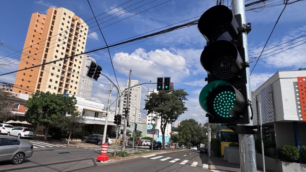 A imagem mostra uma cena urbana durante o dia, com um semáforo em destaque à direita, que está verde. Ao fundo, há prédios altos, um deles com fachada laranja e janelas quadradas, e outro mais claro. O céu está azul com algumas nuvens brancas, indicando um dia ensolarado. Há também árvores verdes ao longo da rua e vários fios elétricos cruzando a parte superior da imagem. Na rua, é possível ver uma faixa de pedestres e alguns carros estacionados ou em movimento. A composição transmite a sensação de um cruzamento movimentado em uma cidade.