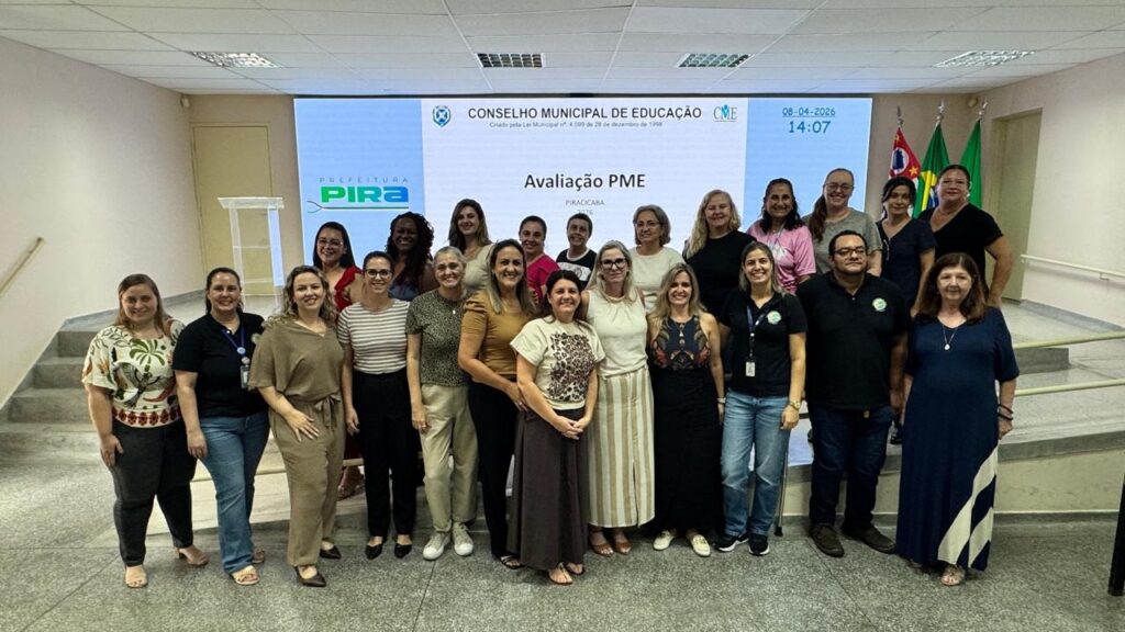 A imagem mostra um grupo de cerca de 19 pessoas, predominantemente mulheres, reunidas para uma foto em um ambiente interno que parece ser uma sala de reuniões ou auditório. Elas estão dispostas em duas fileiras, em frente a uma grande tela digital que exibe o texto "CONSELHO MUNICIPAL DE EDUCAÇÃO", "Avaliação PME", a data "08-04-2024" e a hora "14:07", além do logo da Prefeitura de Piracicaba. Ao lado direito da tela, há três bandeiras: a bandeira do Brasil, a bandeira do estado de São Paulo e a bandeira do município de Piracicaba. O ambiente é bem iluminado, com teto rebaixado e luzes fluorescentes. No canto esquerdo, há um púlpito transparente. As pessoas estão vestidas de forma casual a social, com roupas como blusas, calças jeans, vestidos e sapatos variados. A atmosfera parece profissional e cordial, indicando que o grupo está reunido para uma atividade oficial relacionada à educação, possivelmente uma reunião ou avaliação do Conselho Municipal de Educação de Piracicaba.