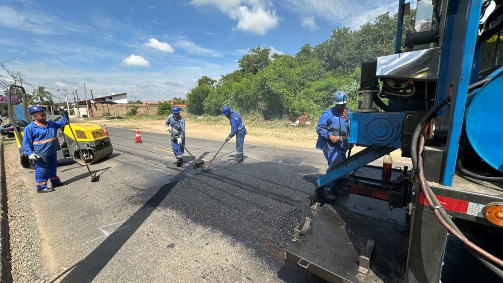A imagem mostra uma equipe de trabalhadores realizando a pavimentação de uma estrada de duas faixas. O cenário é ao ar livre, durante o dia, com céu azul e algumas nuvens. À direita, há uma máquina grande azul usada para espalhar o asfalto quente, e à esquerda, um rolo compactador amarelo está alisando a superfície recém-aplicada. Quatro homens vestindo uniformes azuis, capacetes de segurança e coletes refletivos estão ocupados na tarefa: um opera o rolo, dois nivelam o asfalto com ferramentas manuais, e um está próximo à máquina espalhadora. A estrada está cercada por árvores verdes, sugerindo uma área rural ou suburbana. A luz do sol incide diretamente, criando sombras fortes e destacando as cores vibrantes da cena.