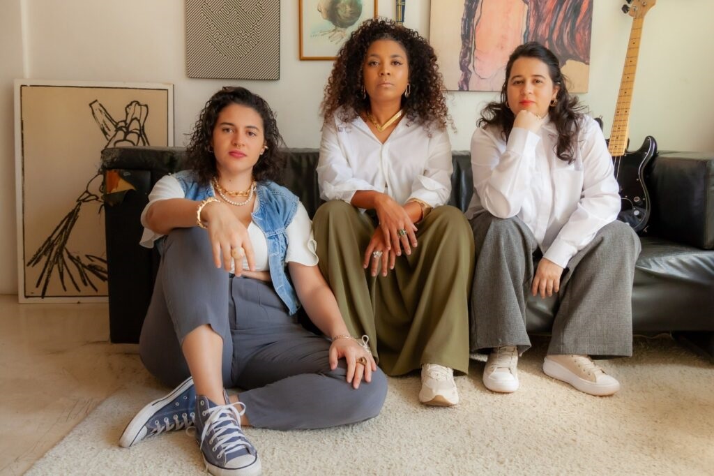 A imagem mostra três mulheres em um ambiente interno, possivelmente uma sala de estar, sentadas e posicionadas de forma descontraída. Elas estão em frente a um sofá preto de couro. A mulher à esquerda está sentada no chão, vestindo um colete jeans sobre uma blusa branca, calça cinza e tênis azul. A mulher no centro está sentada no sofá, vestindo uma camisa branca de botão e calças largas verdes com tênis branco. A mulher à direita também está sentada no sofá, vestindo uma camisa branca de botão e calças largas cinza, com tênis plataforma claro. Todas usam joias douradas, como colares, pulseiras e anéis. Ao fundo, na parede, há quadros decorativos e um violão elétrico preto encostado. A iluminação é natural, vinda da direita, criando sombras suaves e destacando as texturas das roupas e do ambiente. O cenário transmite uma atmosfera calma e elegante, com um estilo moderno e casual.