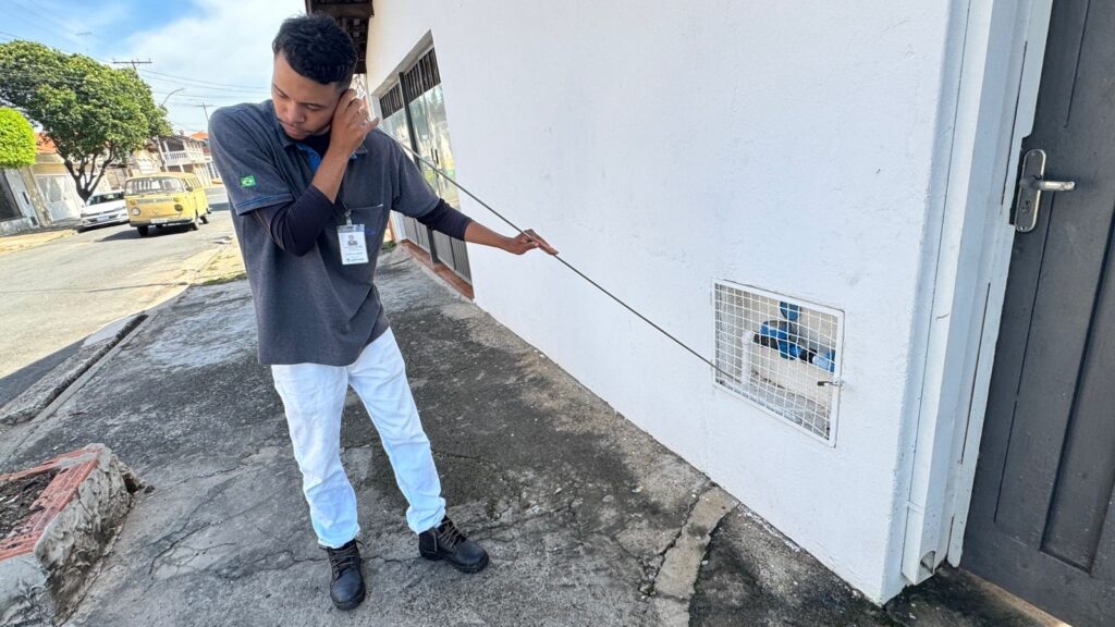 A imagem mostra um jovem vestido com uma camisa polo cinza e calça branca, que está parado em uma calçada ao lado de um prédio branco. Ele segura uma haste longa e fina, que está inserindo em uma pequena abertura gradeada na parede do prédio, onde é possível ver alguns canos azuis. O jovem parece concentrado na tarefa que está realizando. Ele usa uma camisa de manga comprida escura por baixo da camisa polo e tem um crachá preso ao bolso do peito. Ao lado da abertura, há uma porta cinza escura. Ao fundo, é possível ver uma rua com um veículo amarelo, outras casas e árvores, indicando que é uma área residencial ou comercial. A calçada apresenta rachaduras e sinais de desgaste, e há um pequeno canteiro com plantas à esquerda. A iluminação é forte, sugerindo que a foto foi tirada durante o dia, com sombras bem definidas. A cena transmite uma sensação de trabalho ou inspeção relacionada à infraestrutura do prédio.
