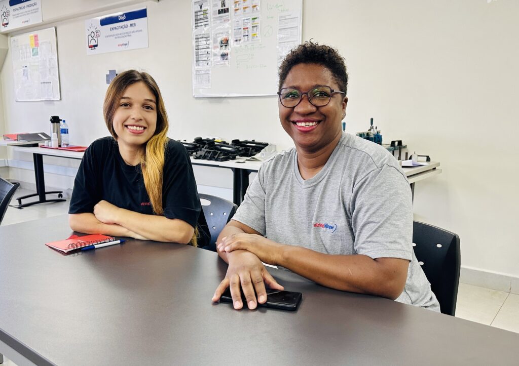 A imagem mostra duas mulheres sentadas lado a lado em uma mesa dentro de um ambiente que parece ser um escritório ou uma área de trabalho. Ambas estão sorrindo e olhando para a câmera. A mulher à esquerda tem cabelo castanho longo e usa uma camiseta preta, enquanto a mulher à direita tem cabelo curto e encaracolado, usa óculos e uma camiseta cinza. Na mesa à frente delas há um caderno vermelho e uma caneta. Ao fundo, há um quadro branco com algumas anotações e diagramas, além de algumas prateleiras com objetos e equipamentos. Na parede, é possível ver placas com textos em português, destacando palavras como "EXPACITAÇÃO-PRODUÇÃO", "Dojo" e frases relacionadas a esforço e resultados. A iluminação é clara e artificial, proporcionando um ambiente bem iluminado e profissional.