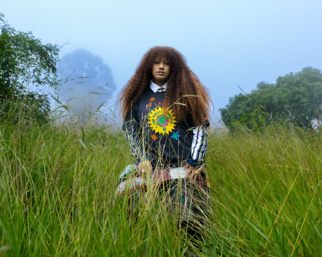 A imagem mostra uma jovem mulher com cabelos castanhos volumosos e cacheados, posicionada em um campo de grama alta e verde. Ela está olhando diretamente para a câmera com uma expressão neutra. Ela veste uma camisa preta de manga longa por cima de uma camisa branca com gola. Na parte frontal da camisa preta, há um desenho colorido que inclui o texto "DE LA" acima de um emblema circular com as palavras "THE MAGIC NUMBER" ao redor e um desenho central que lembra um raio ou estrela. Na parte inferior, ela usa uma peça com padrão colorido, que parece ser uma saia ou um tecido enrolado, com detalhes em branco, rosa e azul, além de franjas coloridas. Ela usa anéis nos dedos e um piercing no nariz. O fundo está desfocado, com árvores e um céu nublado ou enevoado, sugerindo um clima fresco ou úmido. A luz é suave e difusa, típica de um dia nublado, o que valoriza a imagem sem sombras fortes. A composição transmite uma atmosfera serena e levemente mística, reforçada pelo olhar direto da jovem e o cenário natural ao redor.