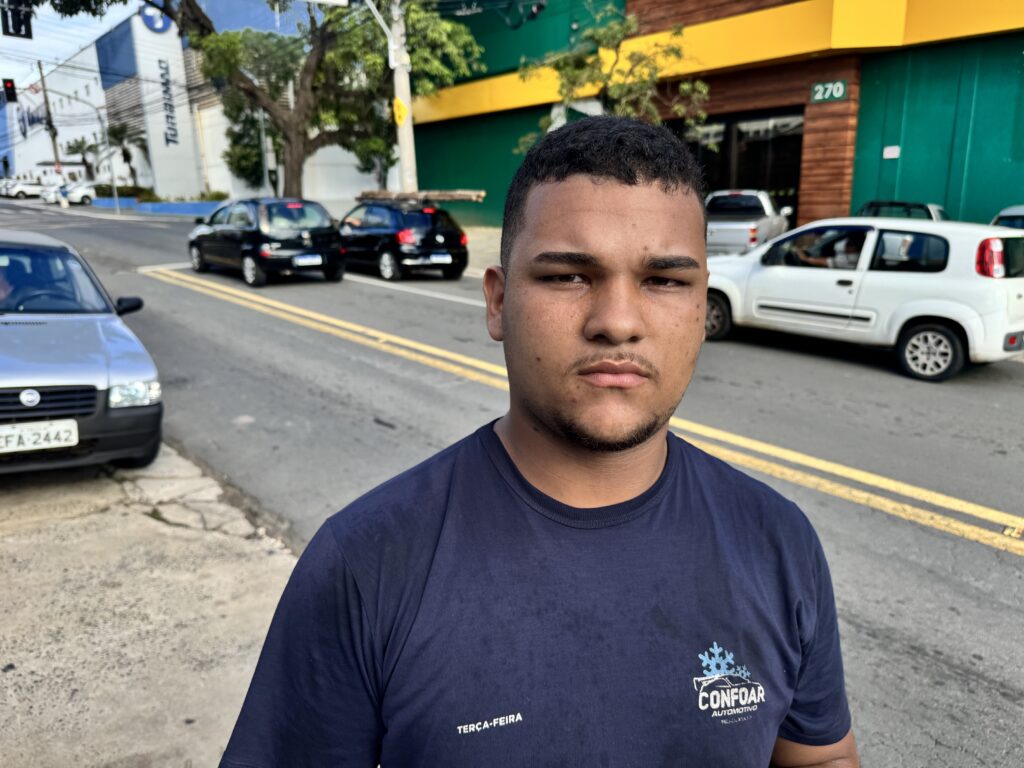 A imagem mostra um homem jovem, de cabelos escuros e barba rala, posicionado em primeiro plano com uma expressão séria. Ele veste uma camiseta azul-marinho com o texto "TERÇA-FEIRA" no lado direito do peito e um logotipo da "CONFOAR AUTOMOTIVO" no lado esquerdo, que inclui um ícone de floco de neve, sugerindo que a empresa atua no ramo automotivo, possivelmente com ar-condicionado ou refrigeração. Ao fundo, há uma rua urbana durante o dia, onde se veem vários carros estacionados, incluindo um Fiat prata com a placa "EFA-2442". Também é possível ver um prédio comercial pintado de verde e amarelo, com o número 270 visível na fachada. A composição está nítida no homem, enquanto o fundo está levemente desfocado, destacando o sujeito principal. A cena parece ter sido capturada em um ambiente de trabalho ou próximo a ele, possivelmente em uma oficina ou loja automotiva no Brasil, dado o idioma português e o estilo das placas dos carros.