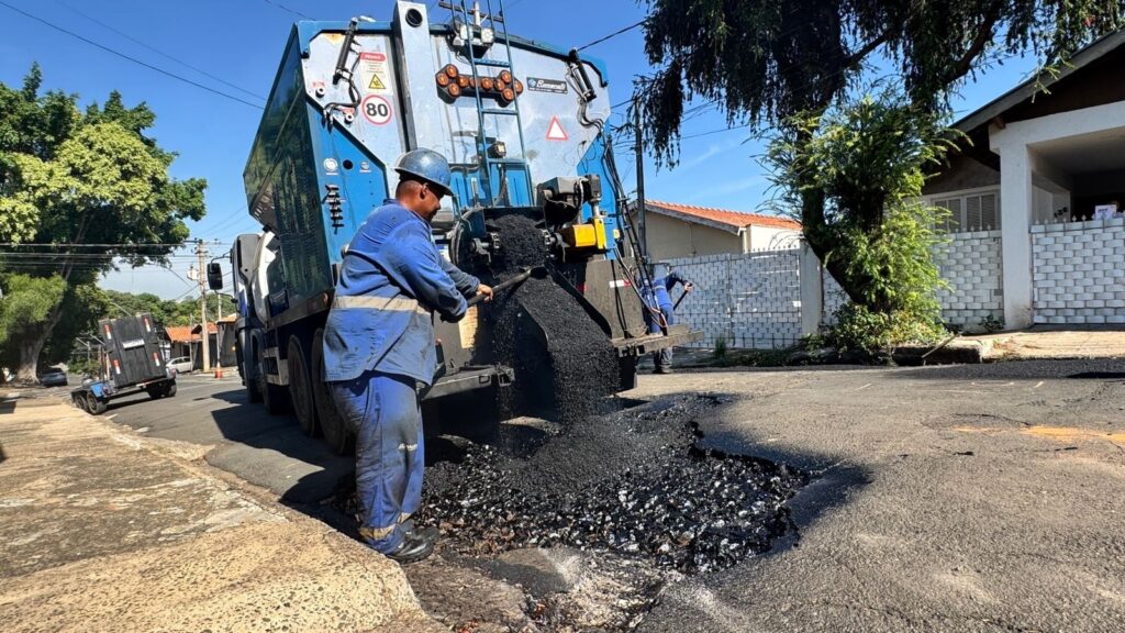 A imagem mostra uma cena de reparo em uma rua durante um dia ensolarado. No centro da imagem, há um caminhão azul especializado em pavimentação, despejando asfalto quente em um grande buraco na pista. Dois trabalhadores, vestidos com uniformes azuis e capacetes, estão auxiliando no processo: um deles utiliza uma pá para espalhar o asfalto dentro do buraco, enquanto o outro está próximo ao caminhão. O buraco na via é bastante evidente, mostrando danos consideráveis no asfalto. O entorno é uma rua residencial, com casas ao fundo, cercadas por muros de tijolos brancos e telhados de cerâmica. Há também uma árvore grande com folhagem verde densa que aparece do lado direito da imagem. O céu está limpo e azul, indicando um dia claro. No caminhão, há placas de sinalização, incluindo uma que indica "PERIGO" e outra com o número "80", provavelmente indicando o limite de velocidade. A marca "Romarali" está visível no caminhão. A imagem foi tirada de um ângulo baixo, destacando o tamanho do caminhão e o buraco na rua. Em resumo, a foto retrata uma operação de manutenção viária, onde trabalhadores estão consertando um buraco na rua em um bairro residencial.