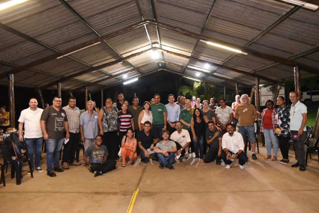 A imagem mostra um grande grupo de pessoas, aproximadamente entre 30 a 40 indivíduos, posando para uma fotografia sob uma estrutura coberta ao ar livre, que parece ser um pavilhão ou um abrigo aberto com teto de metal ondulado sustentado por vigas e pilares de madeira. A iluminação é mista, com luz artificial proveniente de lâmpadas fluorescentes e alguma luz ambiente, criando um ambiente levemente escuro, porém funcional. O grupo está organizado em várias fileiras, com algumas pessoas em pé e outras agachadas ou sentadas na frente. As pessoas são diversas em termos de idade e gênero, todas olhando para a câmera, com expressões que variam entre neutras e sorrisos. O vestuário é casual, com diferentes estilos e cores, o que sugere um evento ou reunião informal. Ao fundo, além da estrutura, é possível ver folhagens escuras e algumas palmeiras, indicando que o local é ao ar livre, possivelmente em uma região tropical ou subtropical. Algumas cadeiras estão posicionadas nas laterais da imagem, e o chão parece ser de concreto ou terra batida. A foto foi tirada em nível dos olhos, capturando todo o grupo de forma ampla e detalhada.