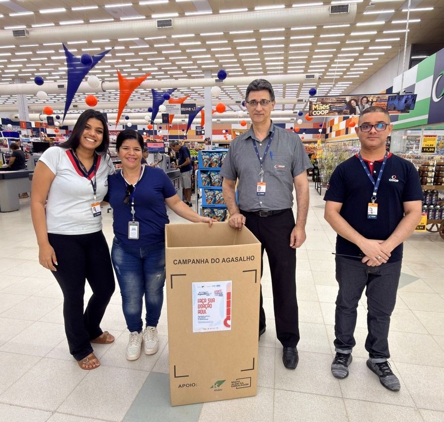 A imagem mostra quatro pessoas em um supermercado, todas usando crachás, provavelmente funcionários ou voluntários. Eles estão em volta de uma grande caixa de papelão destinada à "Campanha do Agasalho", que incentiva as pessoas a fazerem doações de roupas. A caixa tem um cartaz com os dizeres "FAÇA SUA DOAÇÃO AQUI". O ambiente é iluminado e decorado com elementos coloridos pendurados no teto, como triângulos laranja e azul e esferas brancas. As prateleiras ao fundo estão cheias de produtos, típicos de um supermercado. As pessoas estão sorrindo, transmitindo uma atmosfera positiva e colaborativa em relação à campanha. A cena sugere um esforço comunitário para arrecadar roupas para quem precisa durante o inverno.