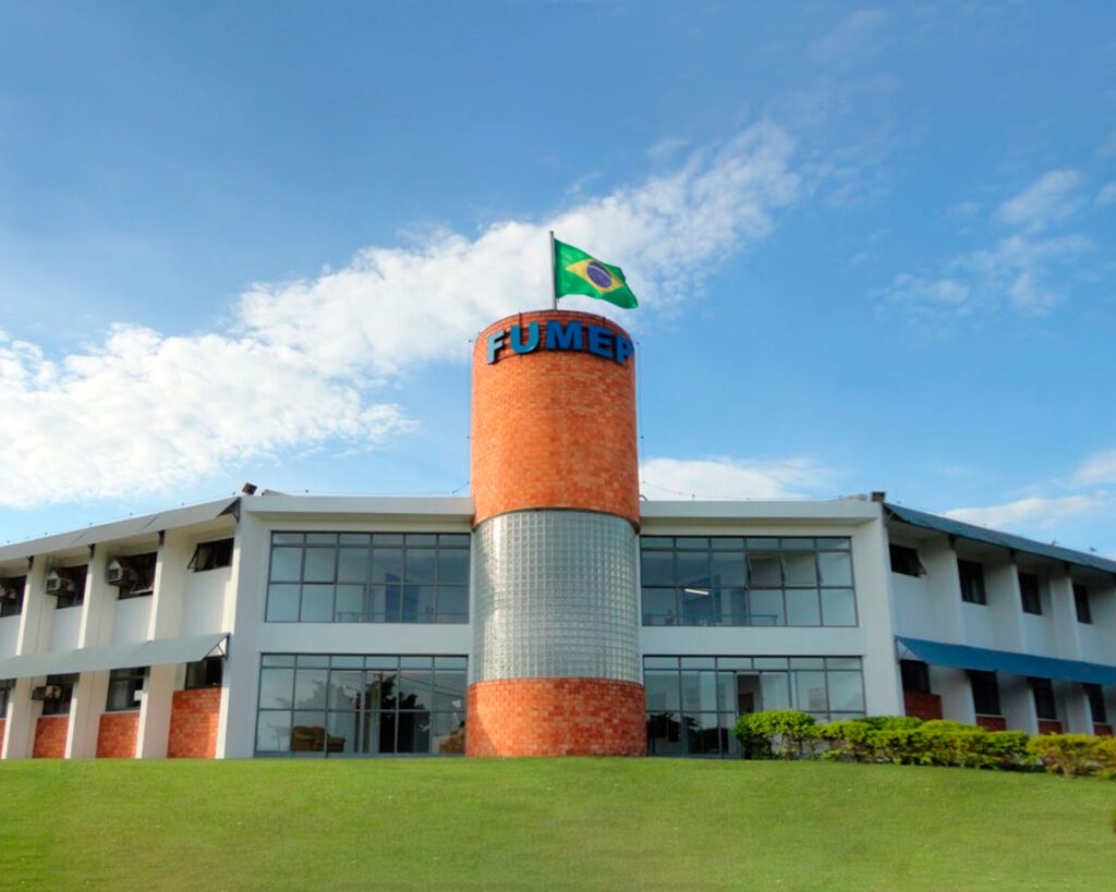 A imagem mostra um prédio moderno de vários andares, possivelmente uma instituição educacional ou administrativa. No centro, destaca-se uma torre cilíndrica feita de tijolos vermelhos com uma faixa de blocos de vidro translúcido no meio, que permite a entrada de luz. No topo dessa torre está a sigla "FUMEP" em letras azuis, e uma bandeira do Brasil está hasteada em um mastro próximo. As alas laterais do prédio são brancas, com grandes janelas de vidro que indicam ambientes internos bem iluminados. A base dessas alas apresenta detalhes em tijolos, combinando com a torre central. Na frente do prédio, há um gramado verde bem cuidado e algumas árvores ou arbustos, o que adiciona um toque natural ao ambiente. O céu está claro, com poucas nuvens, e a iluminação sugere que a foto foi tirada durante o dia, com luz natural. A perspectiva da imagem é de baixo para cima, o que valoriza a altura da torre central e confere imponência ao edifício.