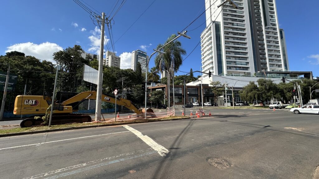 A imagem mostra uma cena urbana em um dia ensolarado, com um céu azul claro e poucas nuvens. No centro-esquerda, há uma grande escavadeira amarela da marca Caterpillar (CAT) posicionada sobre um canteiro de grama, próxima a uma cerca de segurança laranja que delimita a área de obra. No chão, cones de trânsito laranja organizam o espaço ao redor da escavadeira. À direita, destaca-se um edifício alto e moderno, branco, com muitas janelas, que contrasta com outras construções mais baixas ao redor. Ao fundo, é possível ver prédios residenciais altos e algumas árvores verdes que equilibram o cenário urbano. Na parte inferior da imagem, observa-se uma rua com algumas marcas de sombra e luz, além de poucos veículos circulando, incluindo um carro branco. O ambiente transmite uma sensação de movimentação e desenvolvimento urbano, com a obra em andamento e a arquitetura contemporânea ao redor. A foto foi tirada de um ângulo baixo, o que valoriza a dimensão do maquinário e dos edifícios.