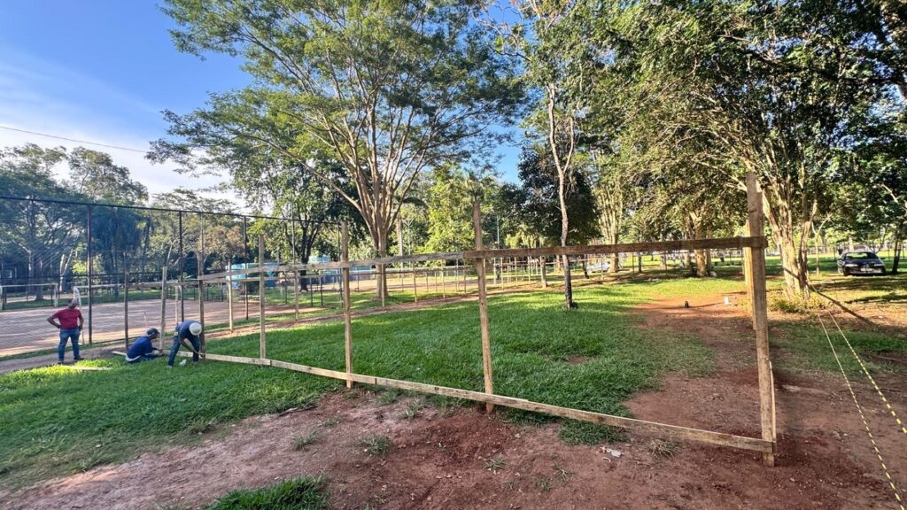 A imagem mostra uma cena ao ar livre, onde está em andamento a construção de uma cerca de madeira. A cerca está parcialmente construída, com postes verticais e vigas horizontais de madeira crua. Alguns postes estão firmemente fixados no chão, enquanto outros ainda estão sendo instalados. No primeiro plano, há três homens trabalhando na construção. Um deles, vestido com camiseta azul e calça escura, está ajoelhado, aparentemente fixando uma viga a um poste. Outro homem, usando capacete branco, camiseta azul e calça escura, também está próximo ao chão, provavelmente martelando ou fixando alguma peça. Um terceiro homem, com camiseta vermelha e jeans, está em pé, observando o trabalho. O ambiente é natural, com grama verde e árvores ao fundo, sob um céu azul claro e ensolarado. No fundo, há uma cerca de arame que delimita uma quadra esportiva, possivelmente uma quadra de tênis, indicada pelas estruturas azuis visíveis. Um carro escuro está estacionado próximo às árvores. A cena transmite a ideia de um trabalho em progresso, com foco na construção da cerca em um espaço ao ar livre, possivelmente em um parque ou área de lazer.