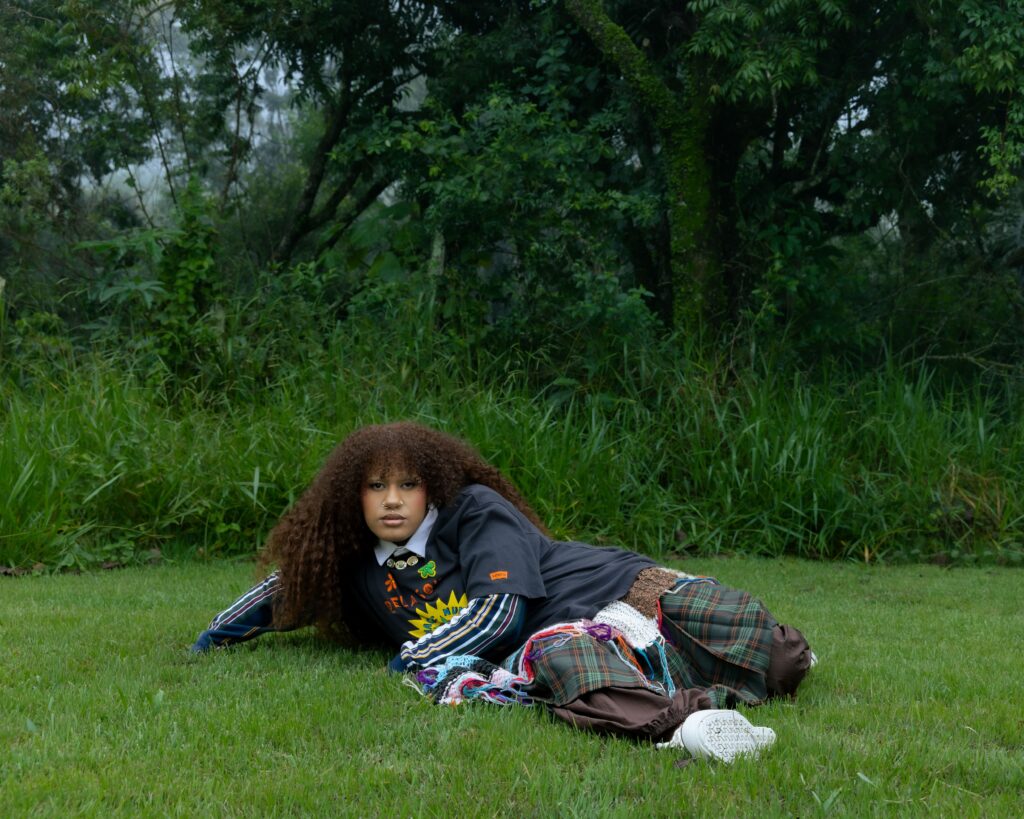 A imagem mostra uma pessoa jovem deitada sobre a grama verde e exuberante. Ela tem cabelo volumoso e cacheado, de cor castanha, e olha diretamente para a câmera com uma expressão neutra. Usa um piercing no nariz e maquiagem discreta. A roupa é composta por várias camadas: uma camiseta cinza escura com uma estampa gráfica que inclui um sol amarelo e letras em laranja, usada por cima de uma blusa de manga longa listrada em azul-marinho, verde, amarelo e branco, e por baixo uma camisa branca com gola. No pescoço, ela usa um colar com vários pingentes metálicos redondos. A parte inferior do corpo está coberta por uma saia xadrez verde escura sobre uma calça marrom. Um cachecol colorido, feito em crochê ou tricô, com fios roxos, azuis, vermelhos e brancos, está sobre o colo e a saia. Um tênis branco aparece próximo ao pé direito. O fundo é composto por vegetação densa e árvores, com um clima nebuloso que confere uma atmosfera suave e um pouco etérea. A iluminação é difusa, típica de um dia nublado ou início da manhã. A composição destaca o estilo único e a pose contemplativa da pessoa, criando uma sensação serena e com um toque de estilo boêmio ou vanguardista. Na manga direita da camiseta há uma pequena etiqueta laranja com o logo da marca Levi's.