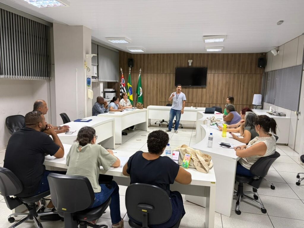A imagem mostra uma reunião ou sessão de treinamento em uma sala de conferências moderna. No centro, um homem de camiseta polo azul clara e jeans está em pé, aparentemente conduzindo a reunião ou apresentando algo para o grupo. Ele usa um crachá no pescoço. Ao redor dele, em formato de U, há várias pessoas sentadas em cadeiras de escritório pretas, prestando atenção ou olhando para seus dispositivos móveis. As pessoas são de diferentes gêneros e estilos, vestidas de forma casual ou casual-esporte. A sala tem paredes brancas, um piso claro e uma parede de madeira ao fundo, onde estão penduradas três bandeiras, incluindo a do Brasil, e uma televisão grande. O ambiente é iluminado por luzes fluorescentes no teto, dando uma iluminação clara e uniforme. Sobre as mesas brancas há objetos como laptops, garrafas de água, cadernos e celulares, indicando que é um ambiente de trabalho ou estudo colaborativo. A disposição em U das mesas favorece a interação entre os participantes e o apresentador.