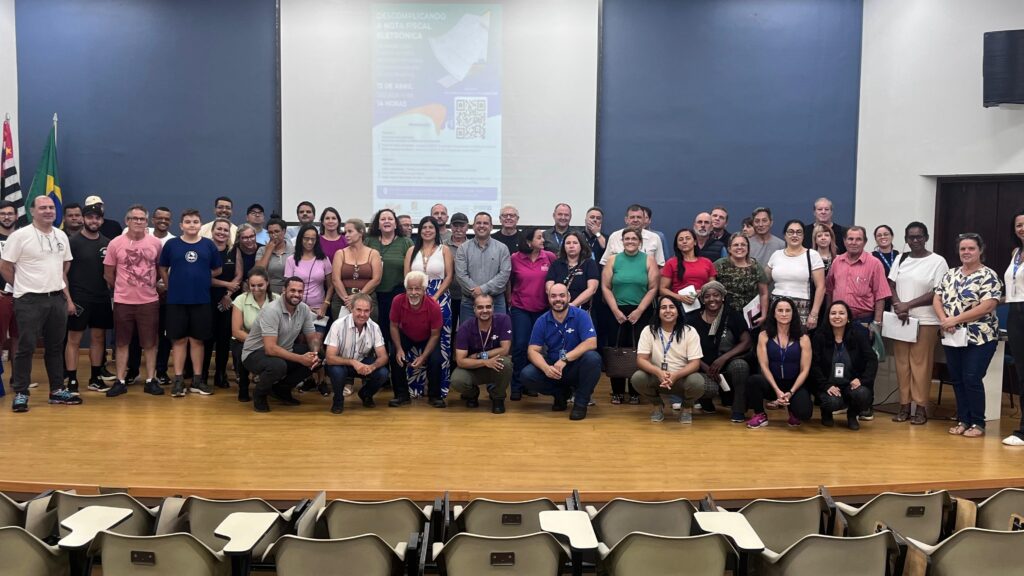 A imagem mostra um grande grupo de pessoas reunidas em um ambiente interno, possivelmente uma sala de conferências ou auditório. Elas estão organizadas em várias fileiras, algumas sentadas em cadeiras dobráveis e outras em pé, todas olhando para a câmera e sorrindo. Ao fundo, há uma parede azul com um telão que exibe um documento com texto e um código QR. O chão é de madeira clara, e há uma bandeira do Brasil à esquerda, indicando que o evento ocorre no Brasil. A iluminação é clara e uniforme, destacando o ambiente e as pessoas. A diversidade do grupo é visível, com diferentes idades e estilos de roupa, que variam entre casual e semi-formal. O clima geral da imagem transmite uma sensação de união, colaboração e celebração, sugerindo que pode ser um encontro, palestra, workshop ou evento comunitário.