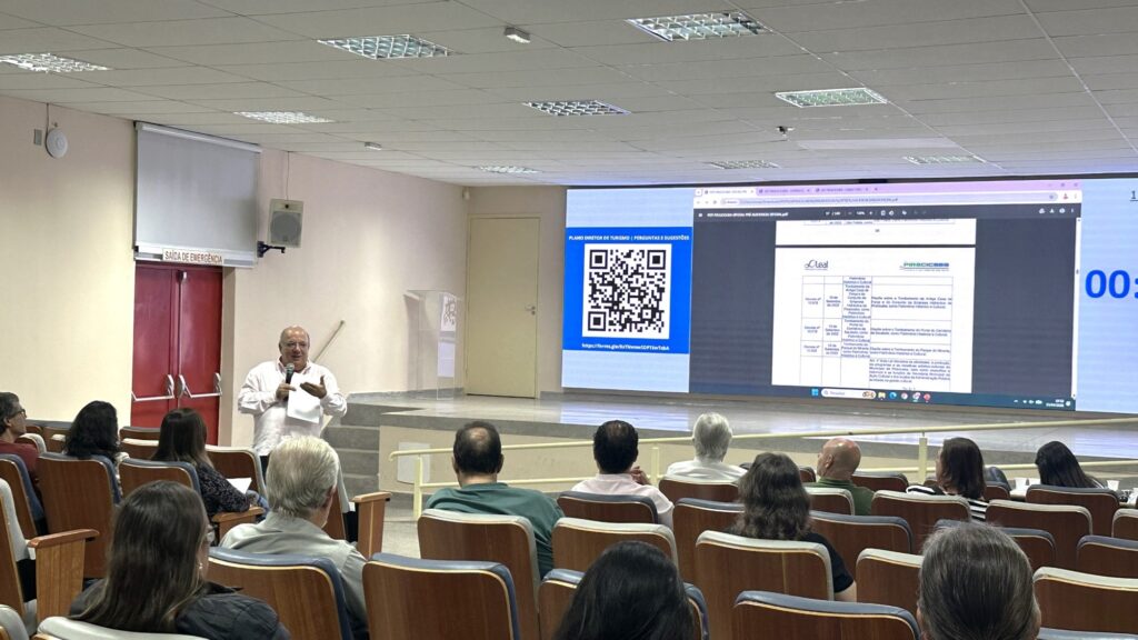 A imagem mostra uma palestra ou apresentação realizada em uma sala de conferências ou auditório. O palestrante, um homem de idade avançada, está na frente, segurando um microfone e um papel, falando para o público. Atrás dele, há uma tela grande exibindo um slide com o título "PLANO DIRETOR DE TURISMO | PERGUNTAS E SUGESTÕES", além de um código QR que provavelmente direciona para um formulário online, indicado pelo link do Google Forms visível na tela. A plateia está sentada em cadeiras alinhadas, com as costas voltadas para a câmera, atenta à apresentação. A sala possui teto com placas acústicas e iluminação fluorescente. As paredes são claras, com portas de emergência vermelhas à esquerda da tela. O ambiente é moderno e funcional, adequado para eventos e reuniões. O foco da apresentação parece ser a coleta de perguntas e sugestões do público sobre o plano diretor de turismo, sugerindo que se trata de uma reunião pública ou consulta para planejamento turístico. A imagem transmite uma atmosfera profissional e organizada, com boa iluminação e clareza visual.