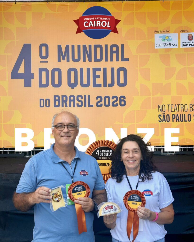 A imagem mostra duas pessoas, um homem e uma mulher, posando em frente a um grande banner laranja com detalhes amarelos, que anuncia o "4º Mundial do Queijo do Brasil 2026". No topo do banner está escrito "Queijos Artesanais Cairol", indicando a marca dos queijos. O homem, vestido com uma camisa polo azul e usando óculos, segura na mão esquerda um pedaço de queijo embalado e, na mão direita, uma fita de prêmio bronze com o logo da Cairol. A mulher, vestindo uma camiseta branca, também segura um pedaço de queijo e uma fita bronze semelhante. Ambos usam crachás pendurados no pescoço, sugerindo que são participantes ou representantes do evento. O banner ao fundo exibe as palavras "Realização: Sertão Bras" e "Apoio: Prefeitura São Paulo", além do local do evento, "No Teatro B São Paulo". A palavra "BRONZE" aparece em destaque sobre a imagem, indicando que eles receberam o prêmio de bronze na competição. A expressão dos dois é de satisfação e orgulho pelo reconhecimento recebido. A iluminação é clara e a foto parece ter sido tirada em um ambiente de evento, possivelmente uma feira ou premiação relacionada a queijos artesanais.