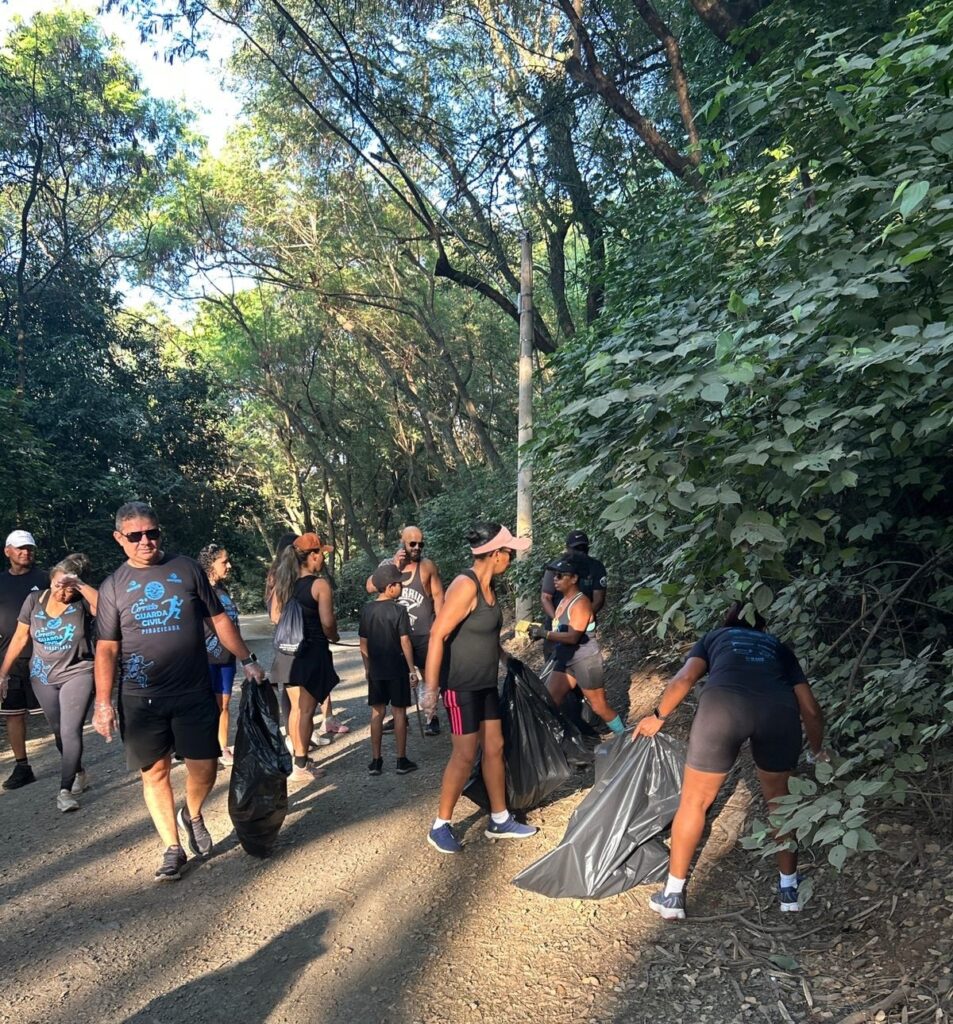 A imagem mostra um grupo de pessoas realizando uma ação de limpeza em uma trilha de terra cercada por vegetação densa. Elas estão recolhendo lixo e colocando em sacos plásticos pretos grandes. A cena ocorre em um ambiente natural, provavelmente um parque ou reserva, com árvores altas e muita sombra, sugerindo que é fim de tarde, com a luz do sol filtrando entre as folhas. As pessoas estão vestidas com roupas casuais e esportivas, demonstrando empenho e concentração na atividade. No fundo, é possível ver mais árvores e um poste de energia. A atmosfera transmite um sentimento de cooperação e cuidado com o meio ambiente.