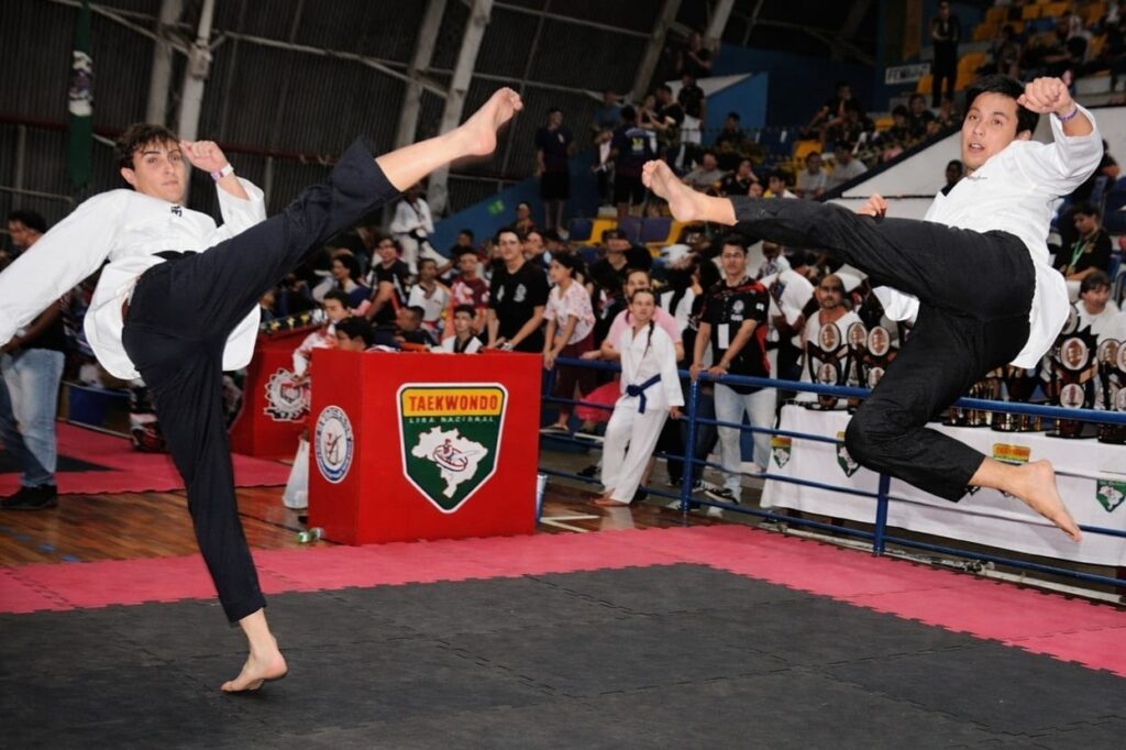 A imagem mostra uma competição de Taekwondo acontecendo em um ginásio esportivo. No centro da cena, dois atletas masculinos estão no ar, executando chutes altos e sincronizados, demonstrando força e técnica. Ambos vestem uniformes tradicionais brancos de Taekwondo, com calças pretas, e estão descalços sobre o tatame preto. Ao fundo, é possível ver uma grande plateia sentada em arquibancadas azuis, assistindo ao combate. À esquerda da imagem, há um estande vermelho com a inscrição "TAEKONDO LIGA NACIONAL" e um logotipo que parece ser o mapa do Brasil, indicando que o evento é uma competição nacional de Taekwondo no Brasil. Próximo ao estande, há uma mesa com troféus expostos, sugerindo que se trata de um campeonato oficial. A iluminação é típica de ambientes internos esportivos, com foco nos competidores, e a fotografia captura o momento exato em que ambos os atletas estão no ar, em pleno movimento, evidenciando a energia e a intensidade da luta. A composição da imagem destaca a ação e também o público presente, mostrando a atmosfera competitiva e o apoio dos espectadores.