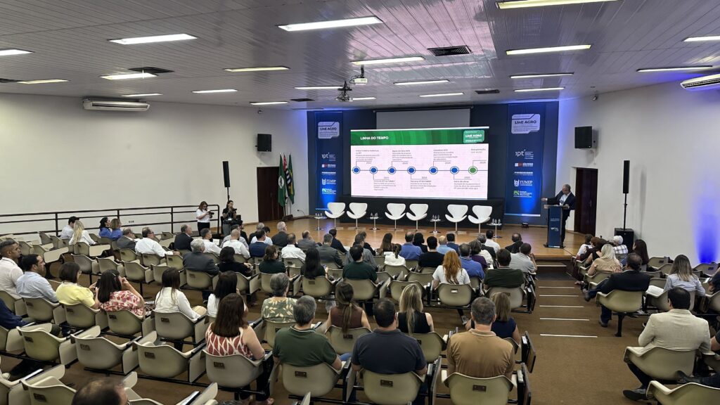 A imagem mostra um auditório grande e moderno durante uma apresentação formal. No centro, há uma tela grande e iluminada exibindo uma linha do tempo intitulada "LINHA DO TEMPO" relacionada ao projeto "LiNE AGRO PIRACICABA". À direita da tela, um homem vestido com terno está falando em um púlpito, provavelmente conduzindo a apresentação. O público, composto por cerca de 60 a 70 pessoas, está sentado em cadeiras alinhadas, prestando atenção ao palestrante. A maioria dos espectadores está usando roupas casuais ou sociais. O ambiente é bem iluminado com luzes de teto embutidas, criando uma atmosfera clara e profissional. No palco, além do palestrante, há seis cadeiras brancas modernas e algumas mesas pequenas, sugerindo que outras pessoas podem participar da apresentação ou debate. À esquerda da tela, dois fotógrafos estão registrando o evento. O fundo do palco é decorado com banners azuis contendo logos de instituições como IPT (Instituto de Pesquisas Tecnológicas), FUMEP, e o Governo do Estado de São Paulo, indicando que o evento tem caráter institucional e está relacionado a parcerias e projetos tecnológicos na cidade de Piracicaba. A linha do tempo exibida apresenta marcos importantes do projeto LiNE AGRO, com datas e eventos desde 2022 até a previsão de inauguração em 2026, destacando visitas, parcerias e início das obras. Em resumo, trata-se de um evento formal que apresenta o desenvolvimento e planejamento do LiNE AGRO, um projeto tecnológico ligado ao setor agroindustrial em Piracicaba, com envolvimento de instituições de pesquisa e governo estadual.