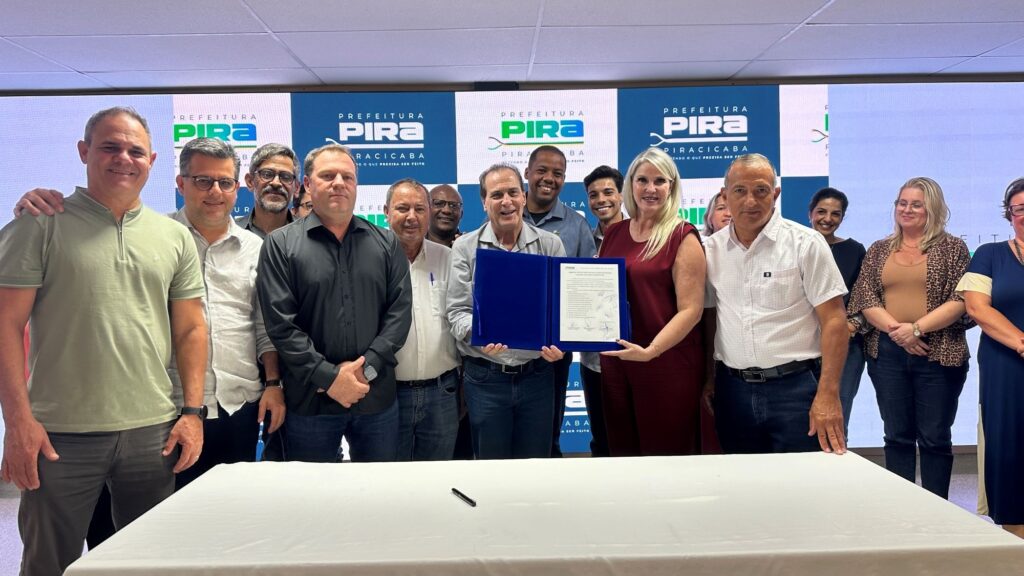 A imagem mostra um grupo de cerca de 14 pessoas reunidas para uma cerimônia formal de assinatura. No centro, um homem com camisa cinza e uma mulher com vestido vinho seguram uma pasta azul aberta que contém um documento assinado, simbolizando a formalização de um acordo ou compromisso. Eles estão atrás de uma mesa coberta com um pano branco, onde há uma caneta preta, provavelmente usada para a assinatura. Ao fundo, há uma grande tela digital com o logotipo da Prefeitura de Piracicaba, repetido em uma grade, junto com o slogan "Fazendo o que precisa ser feito". As pessoas ao redor, de diferentes idades e gêneros, estão sorrindo e olhando para a câmera, demonstrando satisfação e formalidade no evento. Os homens vestem roupas casuais ou sociais, como camisas polo e de botão, e as mulheres usam vestidos ou blusas com calças. A iluminação é clara e uniforme, típica de ambientes internos para eventos oficiais. A composição da foto é ampla, capturando todos os participantes alinhados horizontalmente, evidenciando a importância e o caráter coletivo da ocasião.