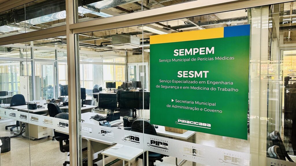 A imagem mostra o interior de um escritório moderno, visto através de uma parede de vidro. Na parede de vidro há uma grande placa verde com texto em amarelo e azul, destacando os nomes "SEMPEM" (Serviço Municipal de Perícias Médicas) e "SESMT" (Serviço Especializado em Engenharia de Segurança e em Medicina do Trabalho). Também aparece a identificação da "Secretaria Municipal de Administração e Governo" e o logo da "Prefeitura de Piracicaba". O escritório é iluminado por luz branca e fria, com um teto industrial onde tubulações e dutos de ventilação ficam expostos. As mesas de trabalho têm computadores, teclados e alguns objetos de escritório, e as cadeiras são ergonômicas e modernas. Na parede de vidro, há um padrão repetido do logo da Prefeitura de Piracicaba em adesivo fosco, criando uma textura visual. Não há pessoas na imagem, e o ambiente transmite uma sensação de organização e funcionalidade, típico de um espaço administrativo público. A composição da foto valoriza a placa verde em primeiro plano, que informa sobre os serviços oferecidos no local, enquanto se vê o escritório ao fundo.
