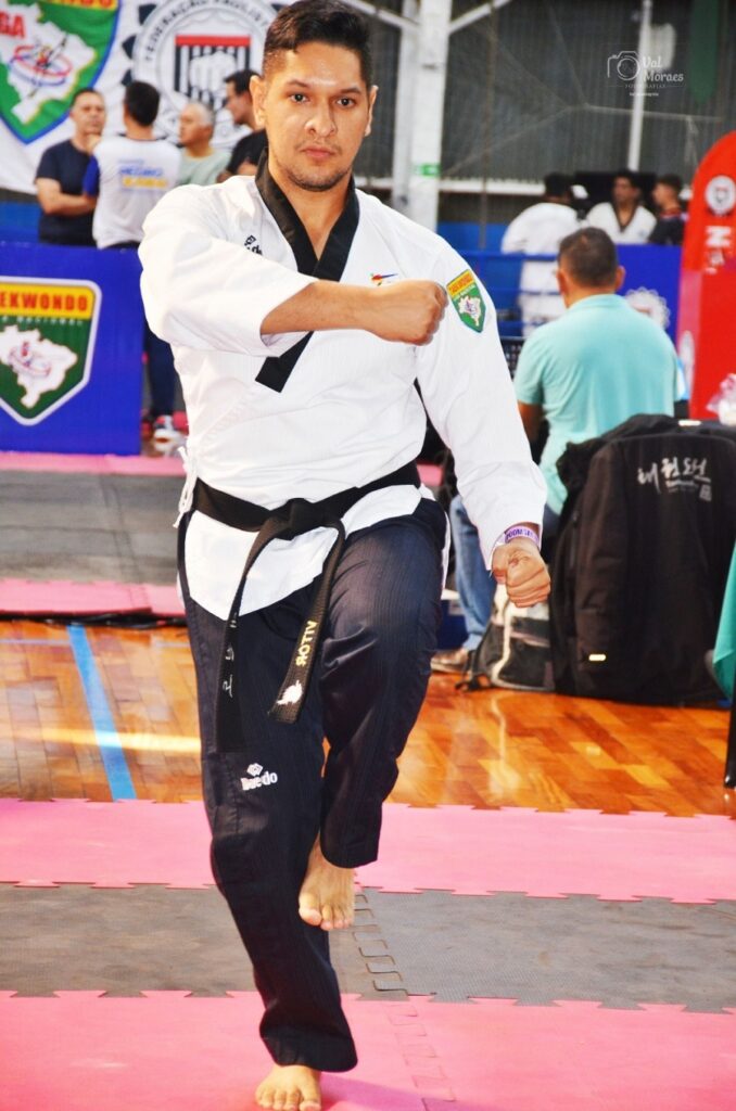 A imagem mostra um homem praticando taekwondo dentro de um ginásio. Ele está vestido com um uniforme tradicional de taekwondo, branco com detalhes pretos, incluindo um cinturão preto indicando um nível avançado. O homem está em uma postura de combate ou defesa, com uma perna levantada e os braços posicionados de forma a proteger o corpo ou preparar um golpe. O fundo revela um ambiente típico de treinamento ou competição, com tatames coloridos (rosa e cinza) no chão e outras pessoas, possivelmente treinadores ou competidores, ao redor. Há também uma faixa azul pendurada com a palavra "TEKWONDO" escrita, além de outros elementos visuais que indicam que o local é uma academia ou espaço dedicado a artes marciais. A iluminação é forte e destaca o atleta, enquanto o fundo está um pouco desfocado, o que ajuda a focar a atenção no praticante. No uniforme, é possível ver algumas inscrições e logotipos, como "Baedo" na calça e um brasão na manga, que provavelmente representam marcas ou associações relacionadas ao esporte. No geral, a imagem transmite concentração, disciplina e a prática do taekwondo em um ambiente profissional e organizado.