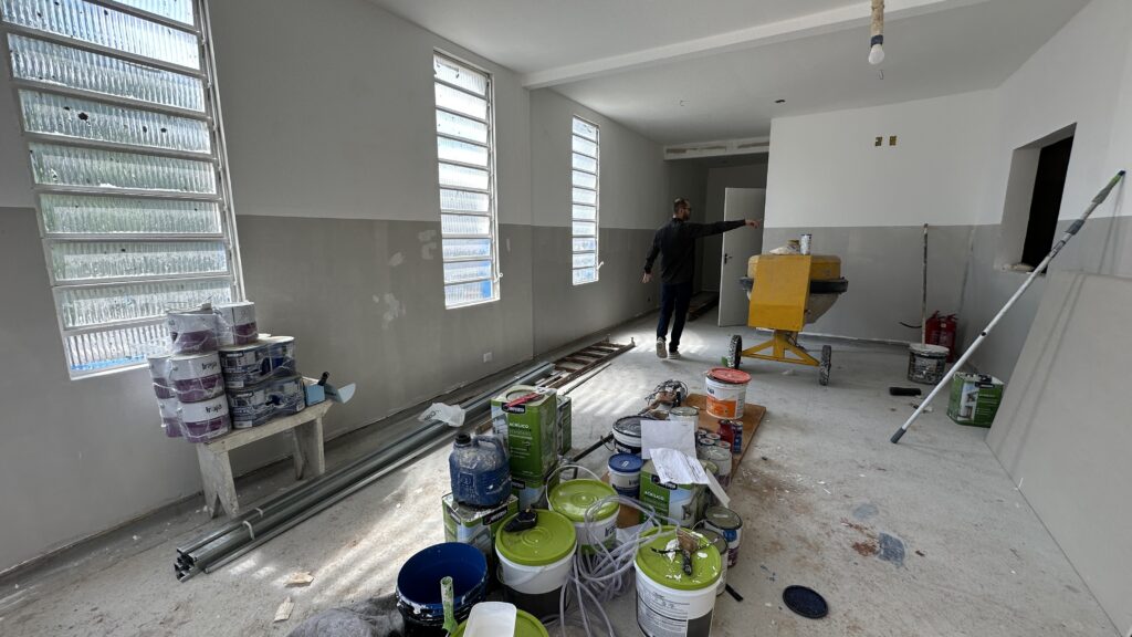A imagem mostra um ambiente interno em processo de reforma ou pintura. É um espaço amplo com várias janelas grandes que permitem a entrada de luz natural. As paredes estão pintadas em duas tonalidades: uma parte superior em tom claro e a parte inferior em azul acinzentado, com algumas áreas ainda em fase de pintura ou preparação. No chão de concreto, há diversos materiais e equipamentos de construção espalhados, como latas de tinta (com a marca "Iraja" e "Universo" visíveis), um misturador de cimento amarelo, pincéis, rolos de pintura, e um extintor de incêndio. Também há tubos metálicos empilhados e um banquinho de madeira apoiando algumas latas de tinta. Um homem veste camiseta e calça escuras e está de costas, apontando para algum ponto à direita da imagem, possivelmente coordenando ou inspecionando o trabalho que está sendo feito. Ao fundo, há portas que dão acesso a outros ambientes. Em resumo, a imagem retrata uma obra ou reforma em andamento, com foco na pintura e preparação das paredes, mostrando o ambiente, os materiais e o trabalhador envolvidos no processo.