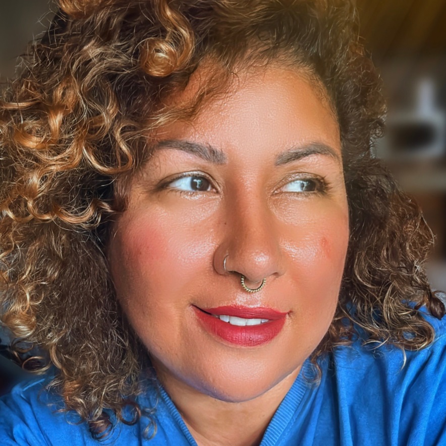 A imagem mostra um retrato próximo de uma mulher com cabelos cacheados e castanhos. Ela está olhando para o lado direito com um leve sorriso no rosto. Ela tem um piercing no septo nasal, com um anel dourado, e está usando batom vermelho. A pele dela tem um tom quente e luminoso, com um leve blush nas bochechas. Ela veste uma blusa azul. O fundo está desfocado, destacando o rosto da mulher. A iluminação é quente e natural, realçando a textura dos cabelos e da pele. A composição foca no rosto e na parte superior do corpo da pessoa.